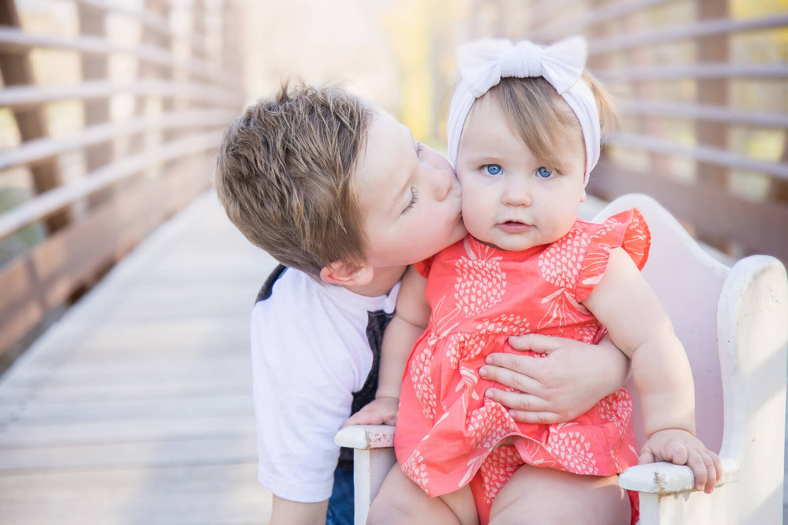 Bright blue eyed toddler girl kissed on the cheek by her cute brother on a bridge