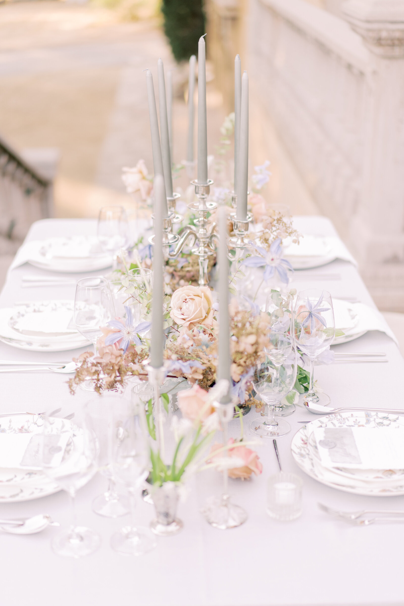 White floral plates, clear glasses, candlesticks, and flowers atop a table with lavender linen outdoors.
