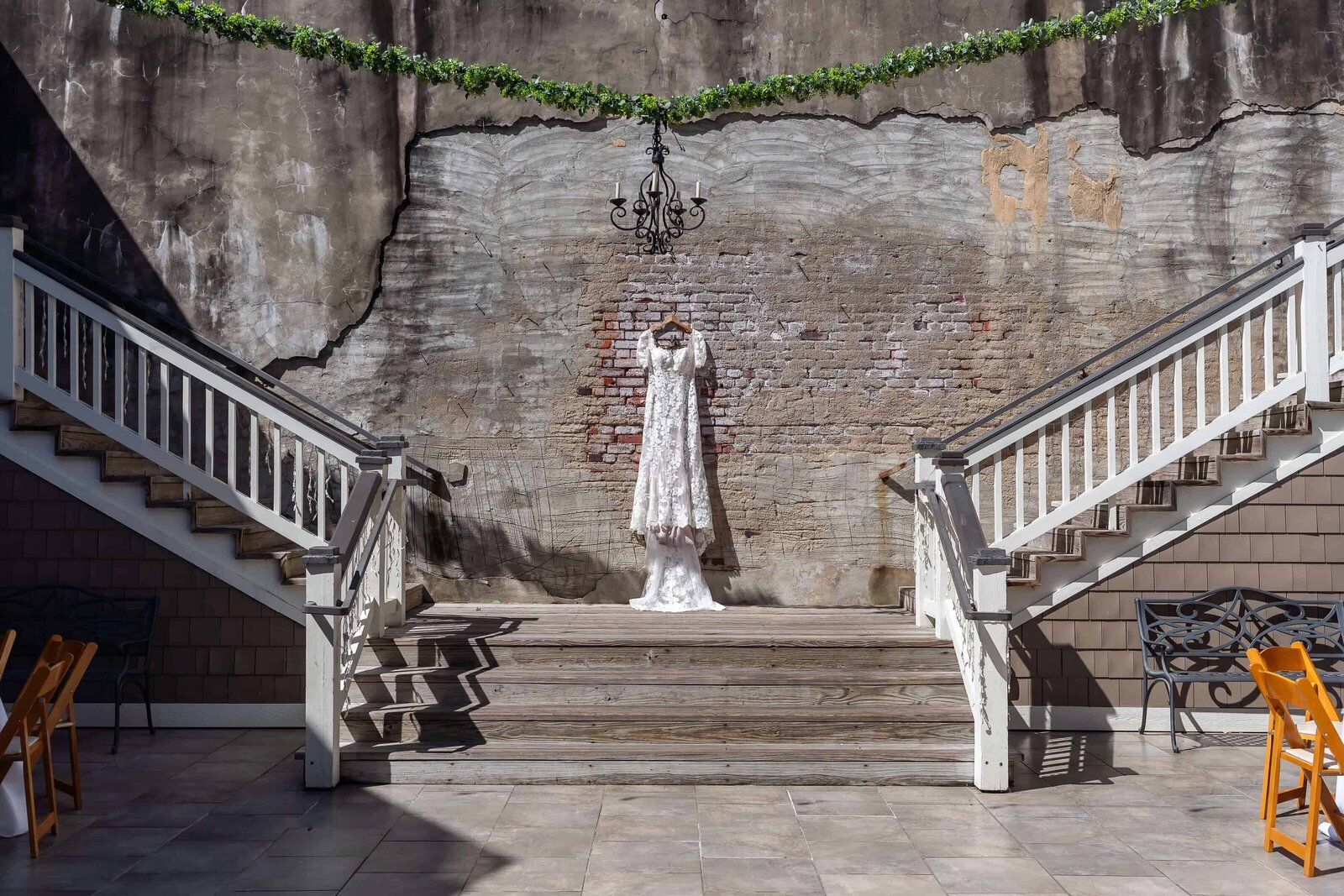 A white lace wedding dress hangs on display between two matching staircases against a weathered brick wall, with a black chandelier and greenery garland above. Sunlight casts clear shadows on the ground.