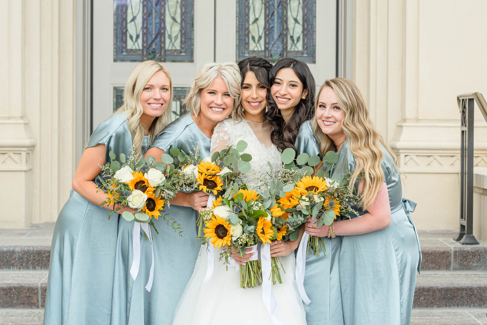 A spring bride and her bridesmaids wearing silky light blue dresses holding bouquets of sunflowers. Captured outside of the Payson Temple in May