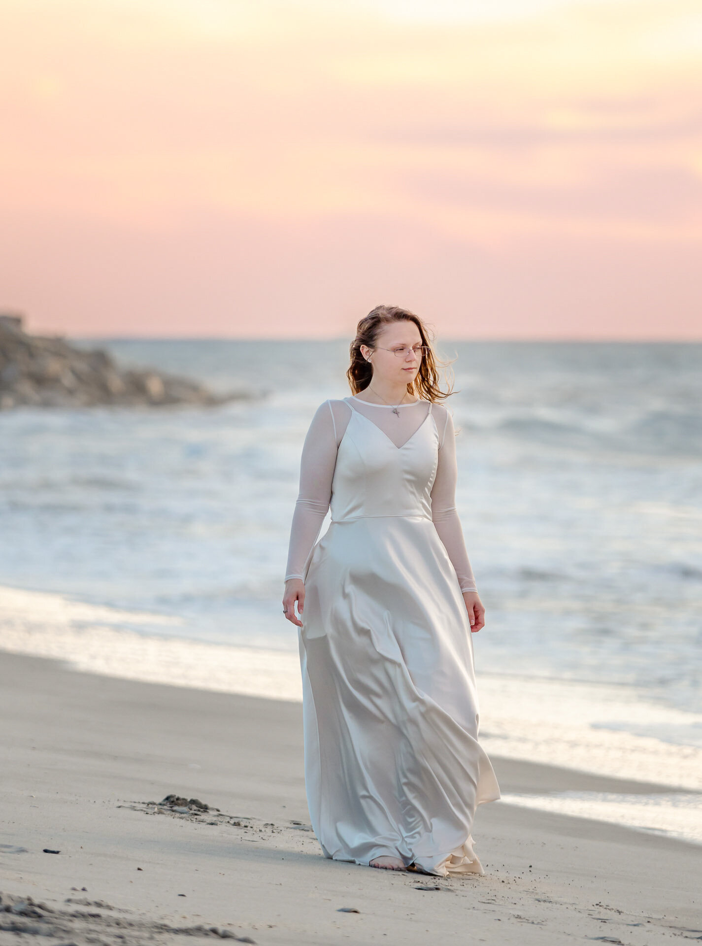 A CFCC senior in a flowing white dress walking along Carolina Beach at sunset near Wilmington, North Carolina. This serene and elegant image captures the beauty of artistic senior photography, ideal for those seeking memorable beach photos.