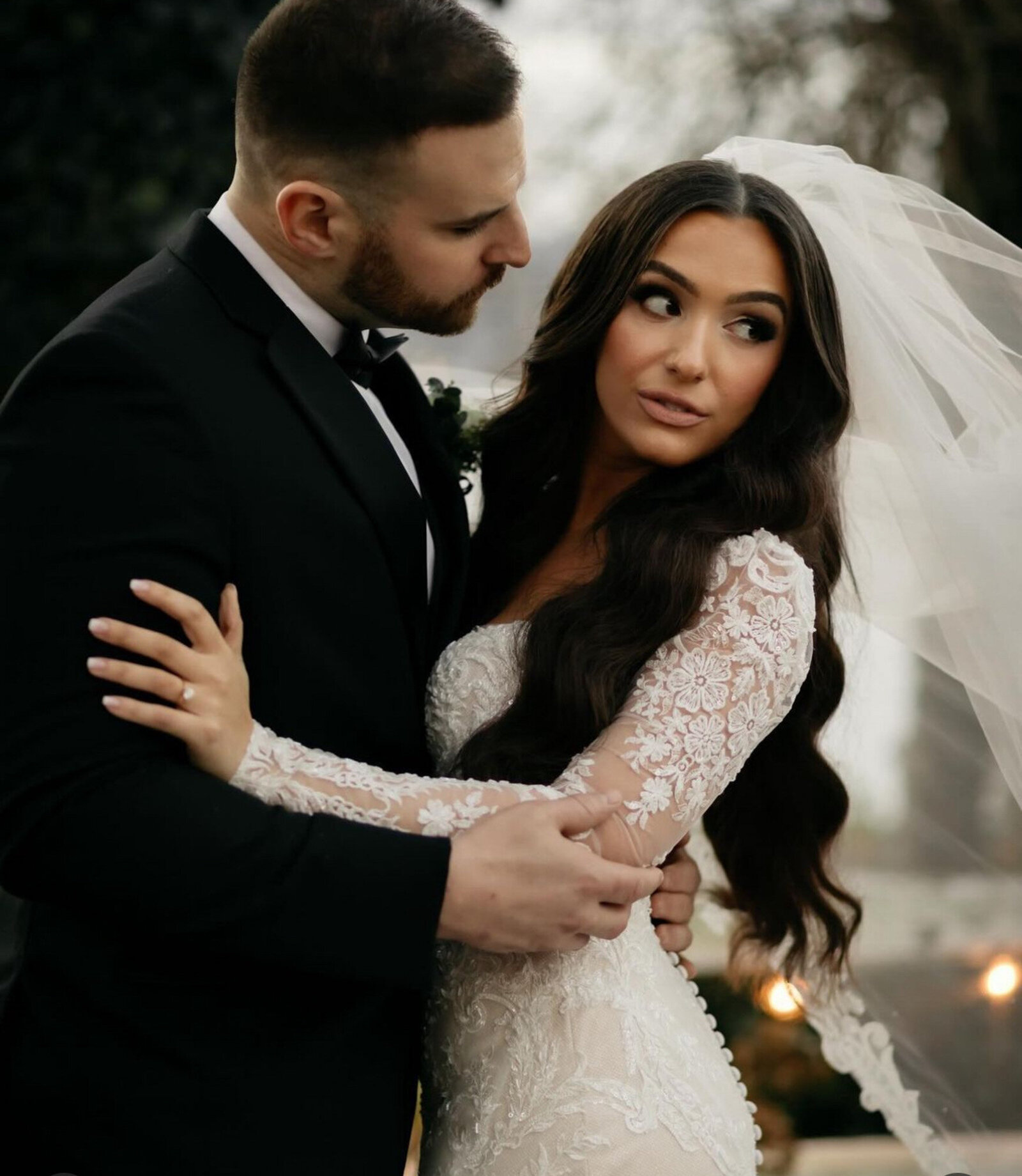 bride wearing a long-sleeve lace wedding dress and veil and groom in a black suit, leans in affectionately towards the bride, embrace each other on their wedding day