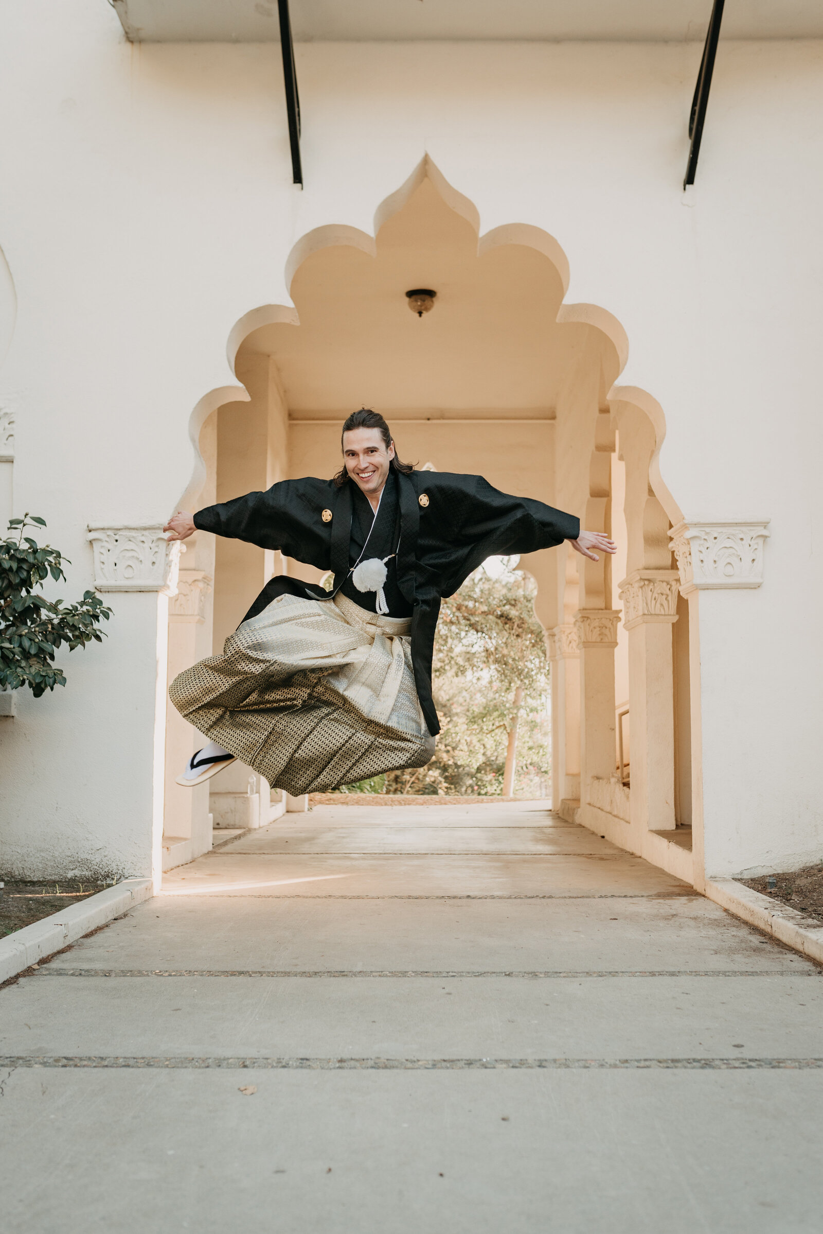 Groom in black montsuki kimono rental jumping with joy under the Moorish-style arch at Brand Park Glendale, Los Angeles California | ブランドパーク グレンデール 着物レンタルウェディング 黒紋付袴でジャンプする新郎