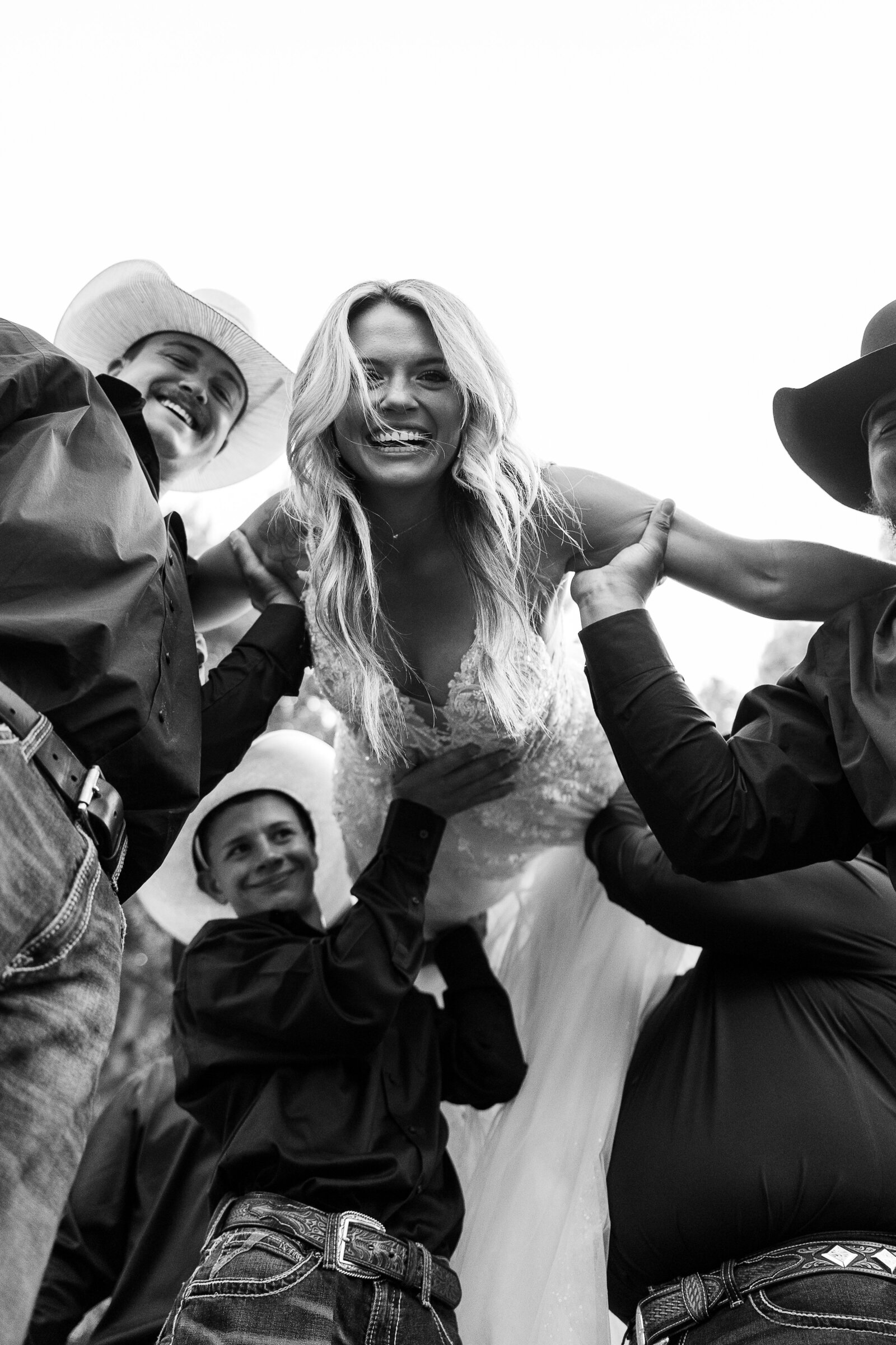 Black and white image of bride being picked up by groomsmen and bride looking down into the camera