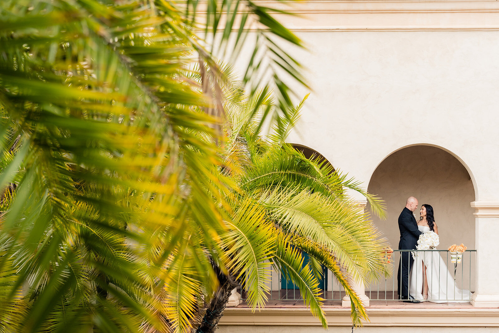 Bride and Groom standing under an archway at The Prado Balboa Park with big Palm Tree leaves in the foreground.