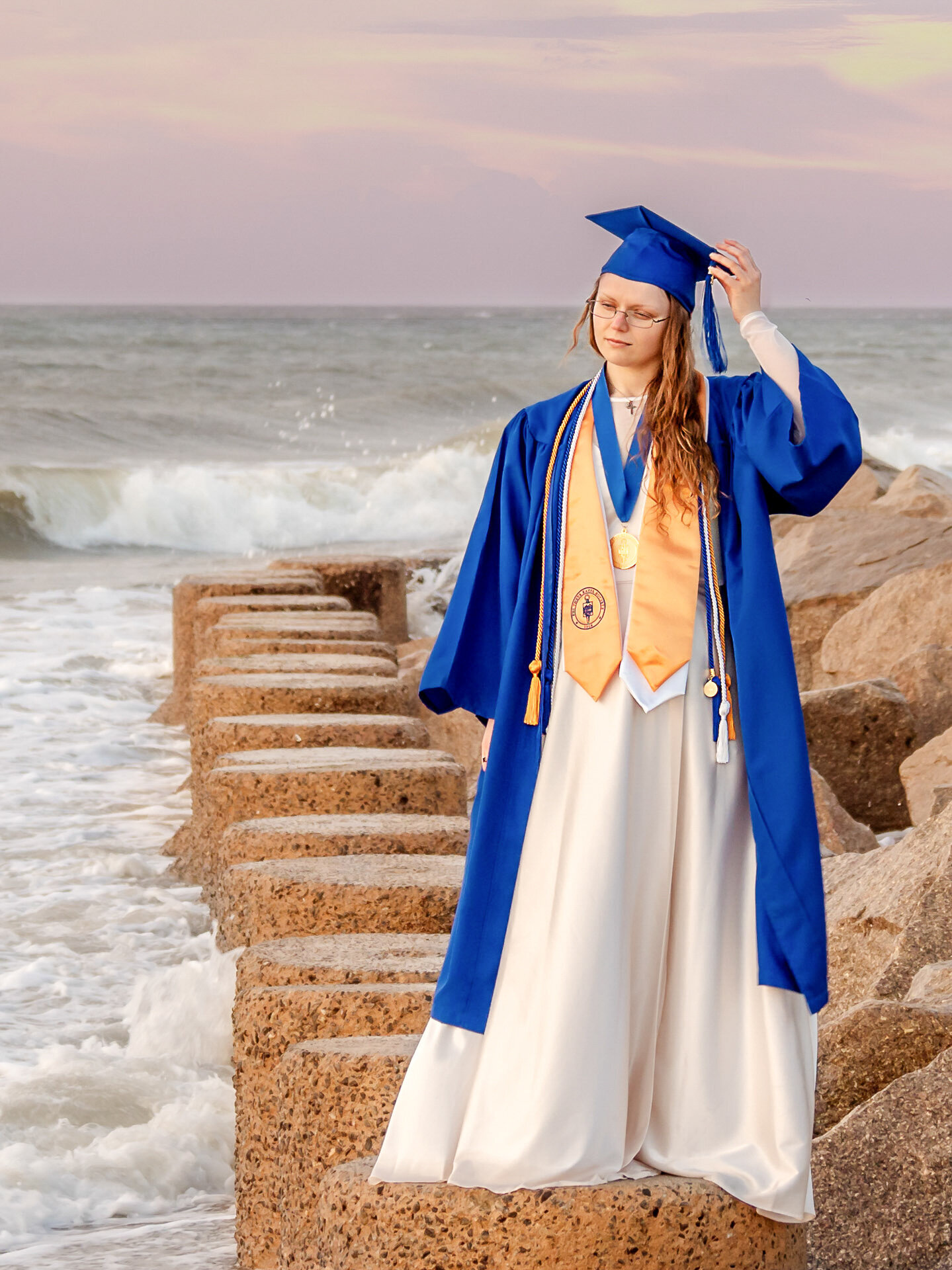 A Cape Fear Community College senior in a blue cap and gown standing confidently on the Fort Fisher beach rocks with waves crashing in North Carolina. This striking photo captures the excitement of graduation, perfect for capturing memorable senior photos.