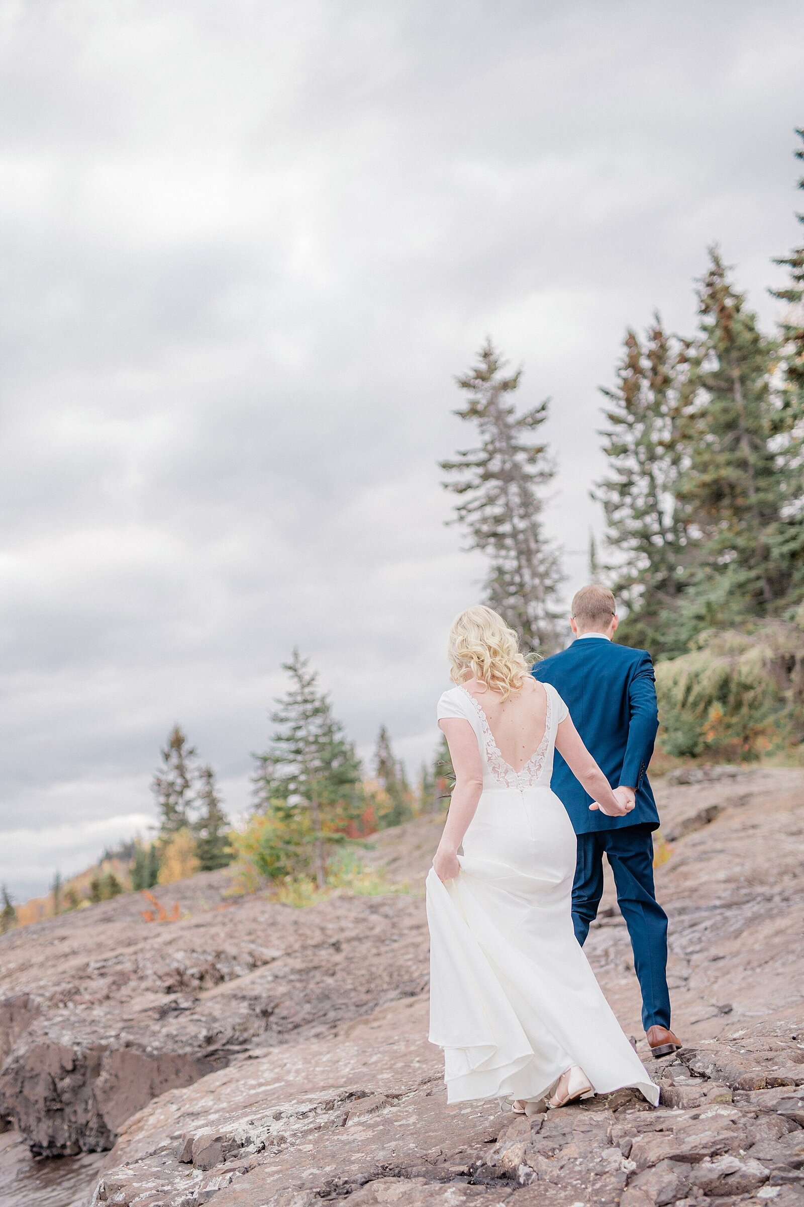 A couple walk away from the camera along the rocks on Lake Superior after their Minnesota wedding.