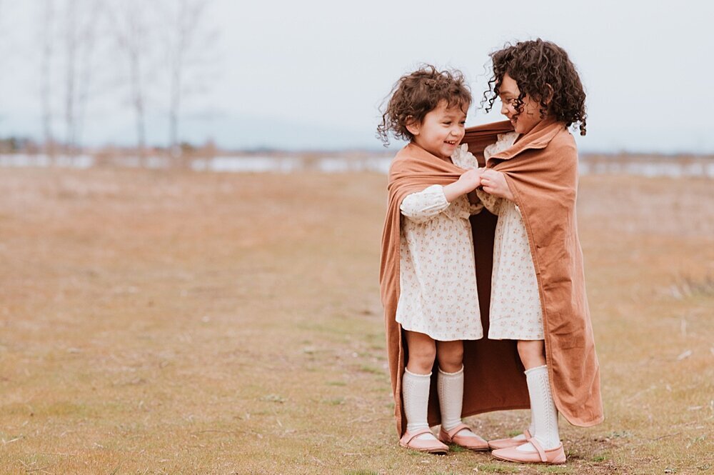 Two sisters snuggling under a blanket in a field with Vancouver Family Photographer