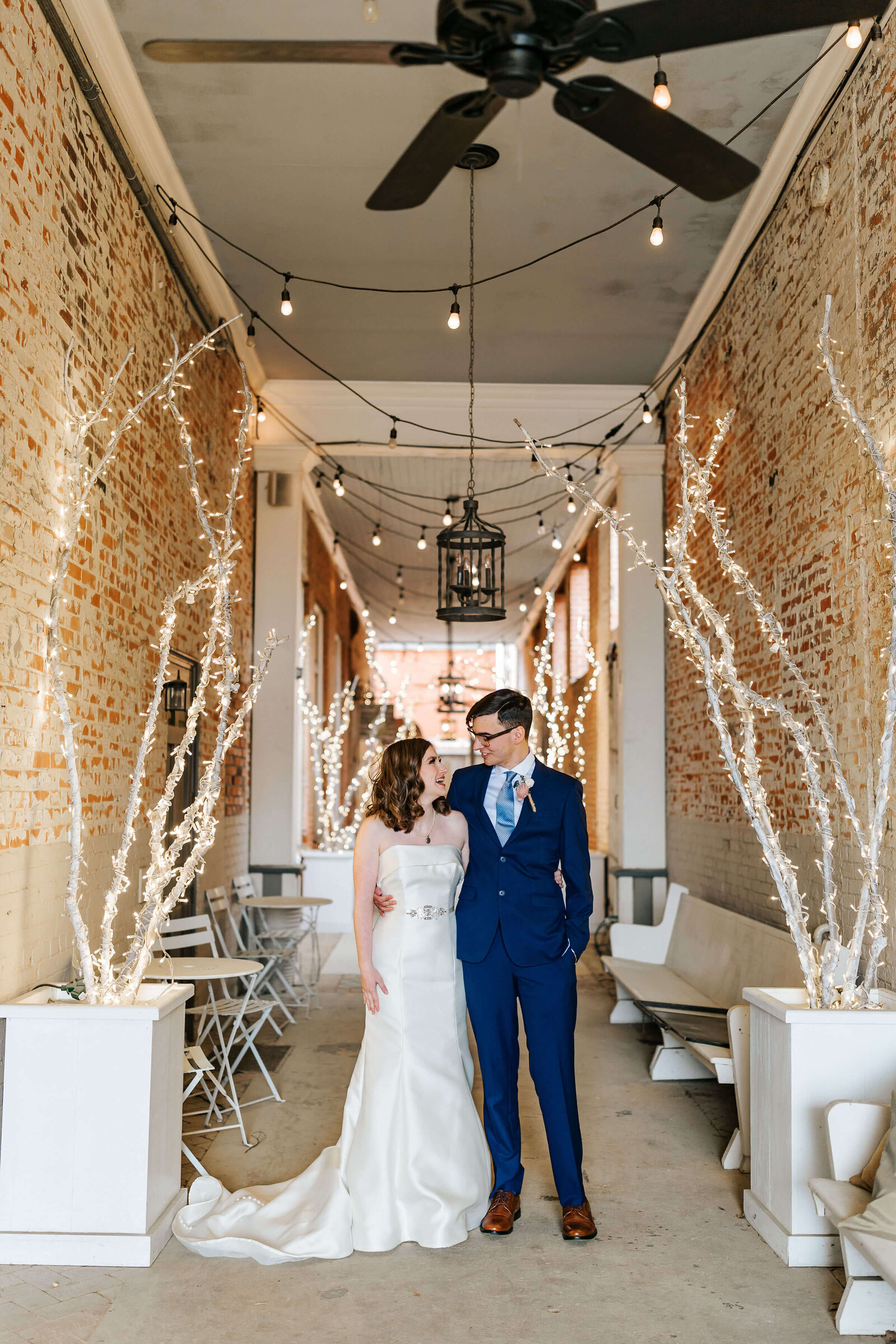 A bride in a white gown and a groom in a blue suit stand together, smiling at each other, in a brick hallway decorated with white lights and bare, illuminated branches.