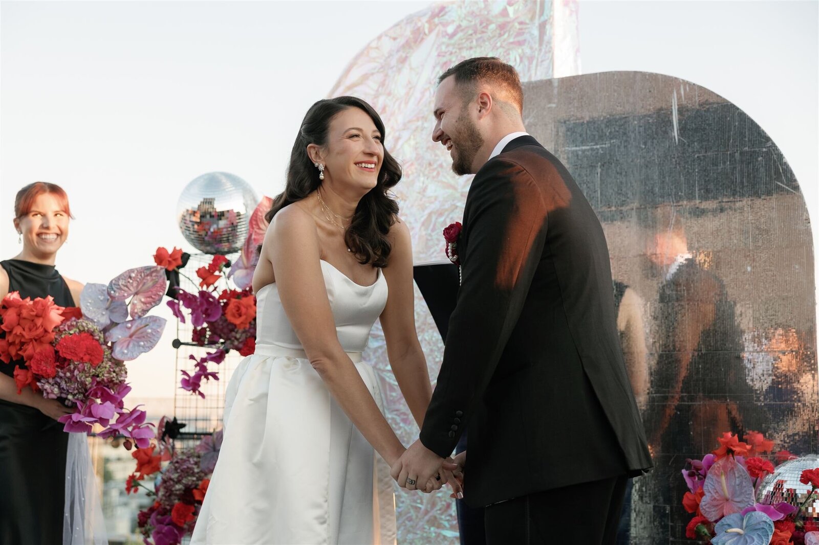 bride and groom at ceremony altar with an iridescent disco inspired backdrop holding hands and laughing