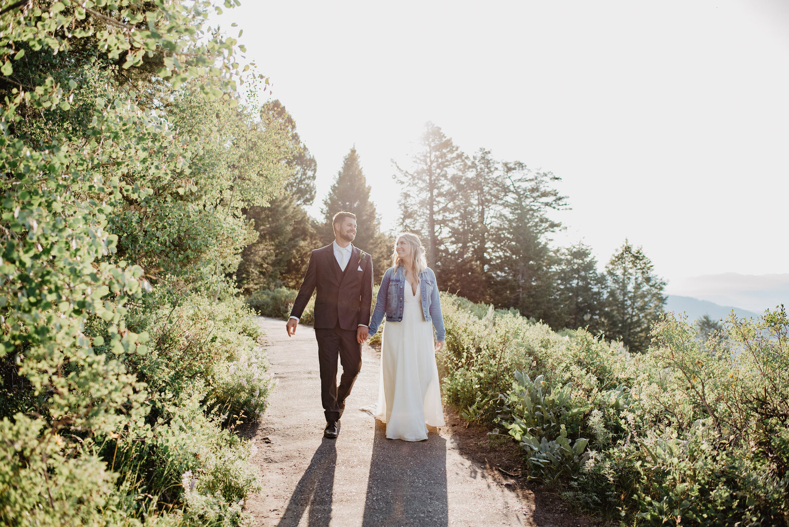 bride adn groom holding hands and walking down a path together in the tetons as they smile at one another while the sun shines from behind them captured by jackson hole wedding  photographers