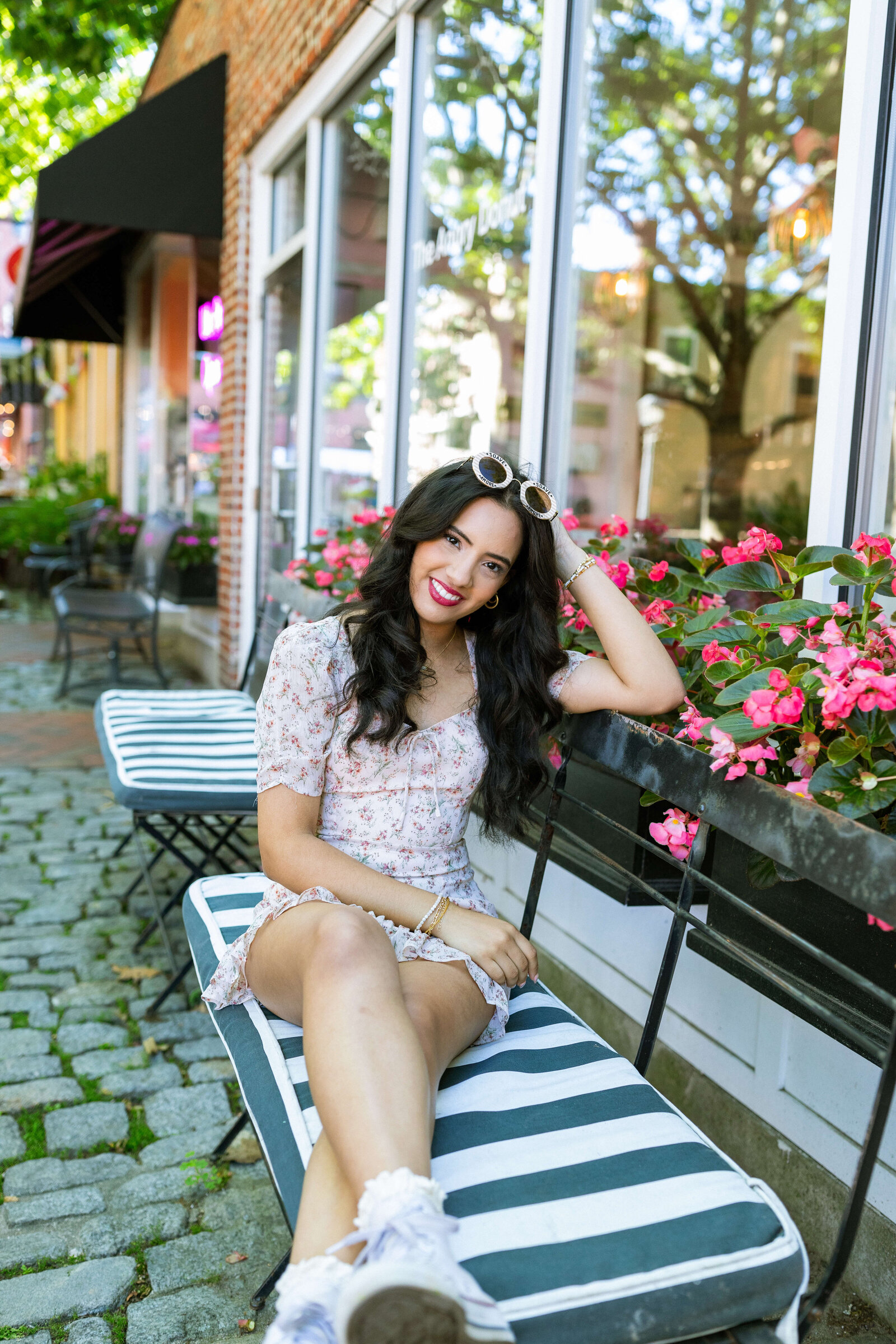 dark haired girl wearing a floral dress with converse and sunglasses sitting on a striped seat in front of flowers