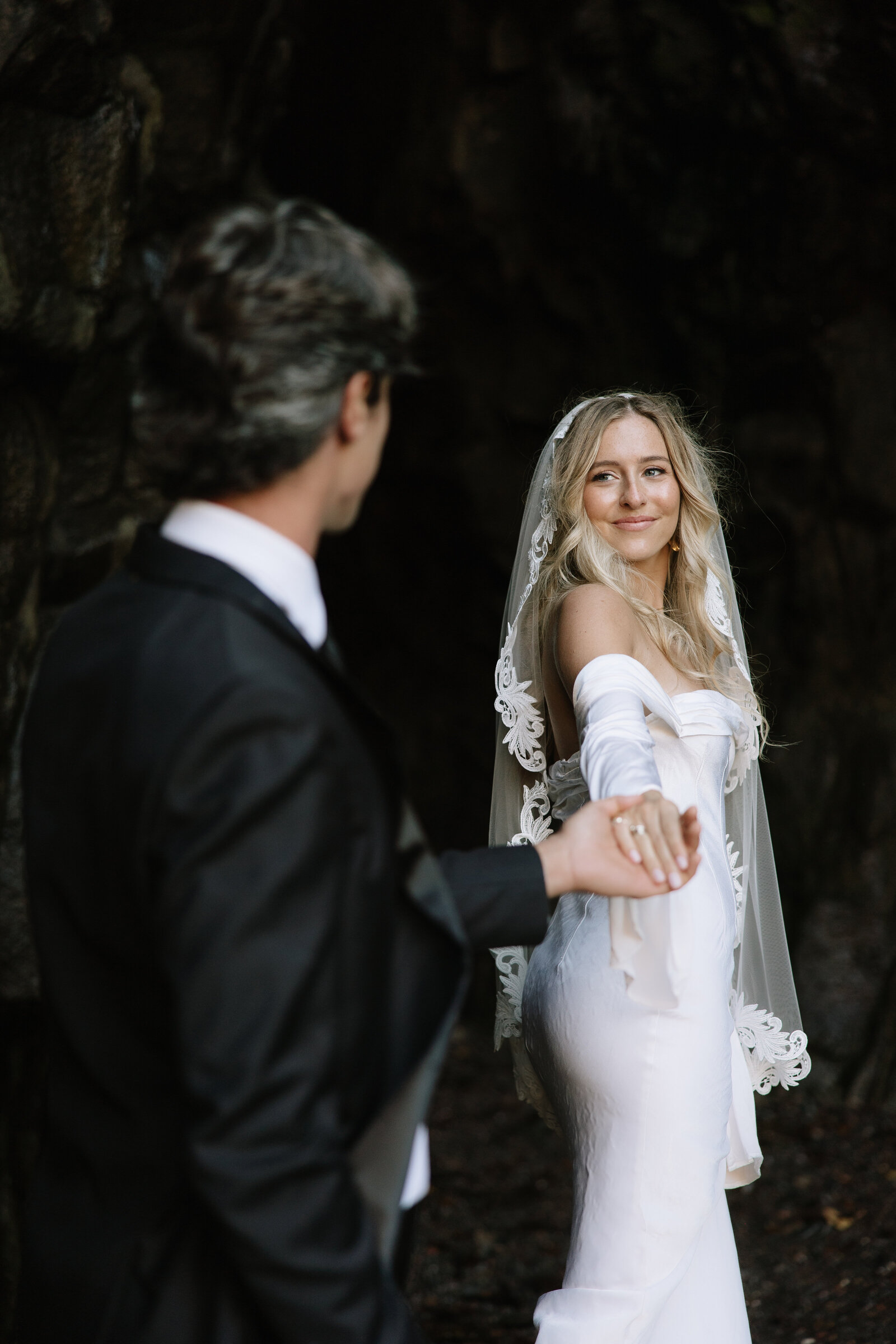A groom holds his wife's hand as she walks across a log in a wedding dress after their elopement. 