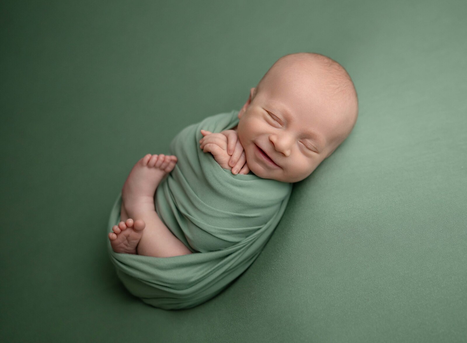 newborn baby boy wrapped in green swaddled and smiling in his sleep. Photo taken at Marietta Newborn photo studio by Jenni Nikki Photography.