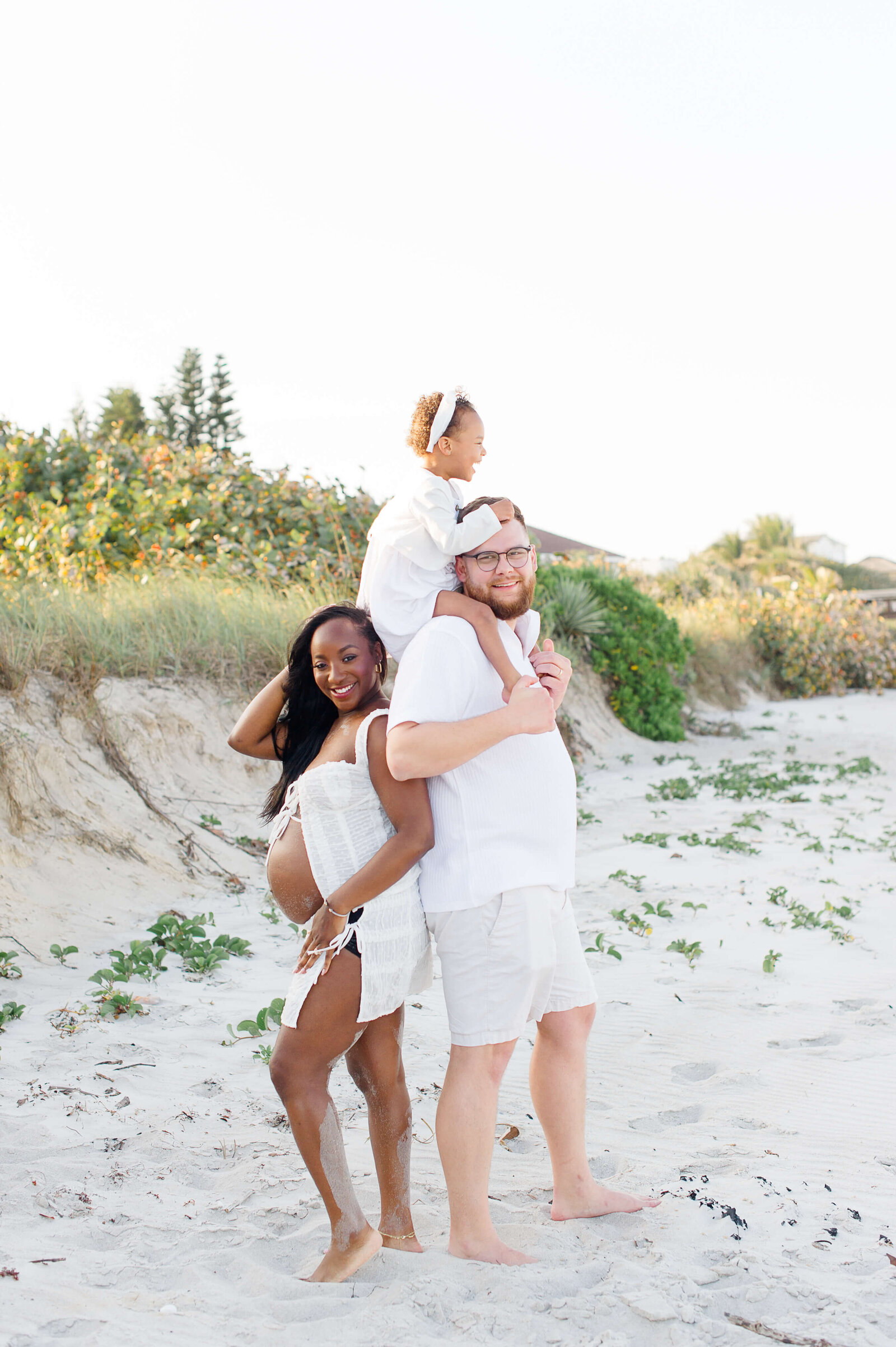 Pregnant parents stand on the beach kissing while daughter plays in the distance