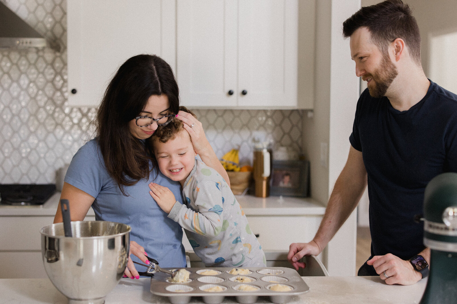 Mom holds son close while dad smiles at the two of them as they are all baking in the kitchen