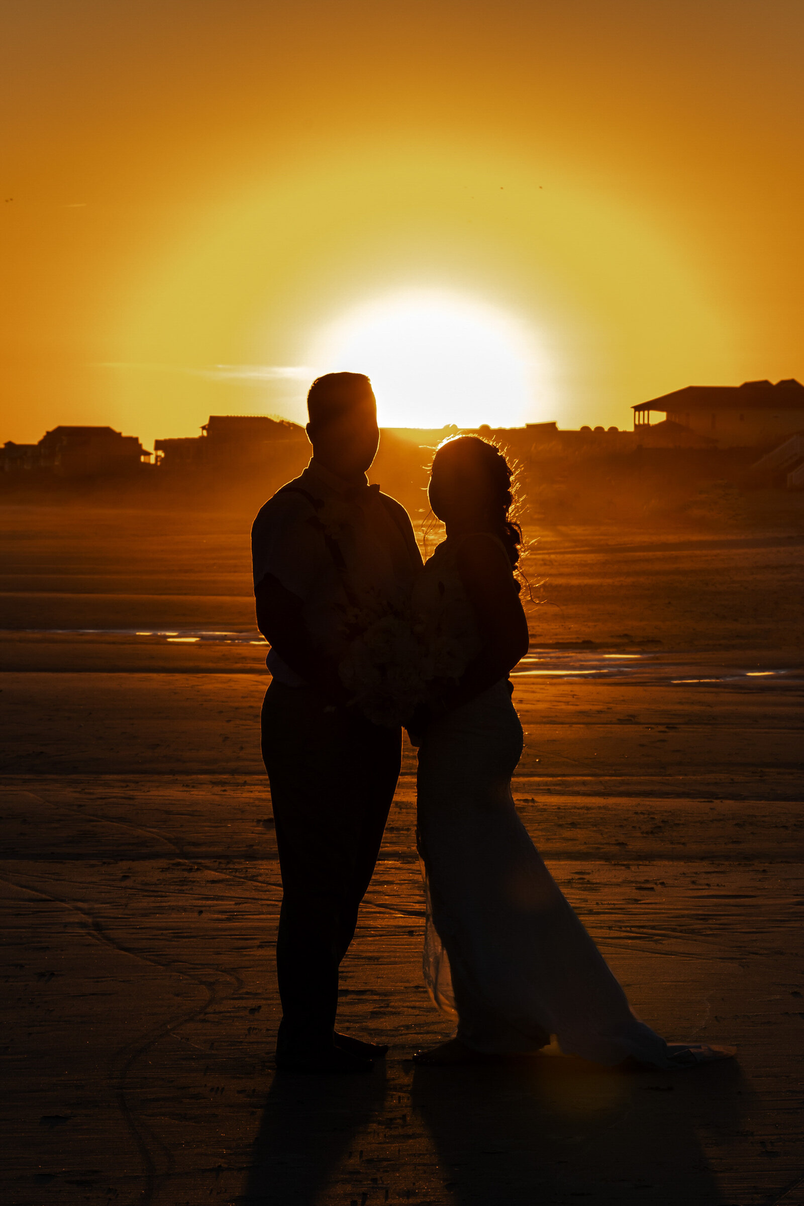 The silhouette of the bride and groom standing on the beach at sunset, with vibrant golden light illuminating the horizon, captured during a romantic Topsail Beach, NC wedding.