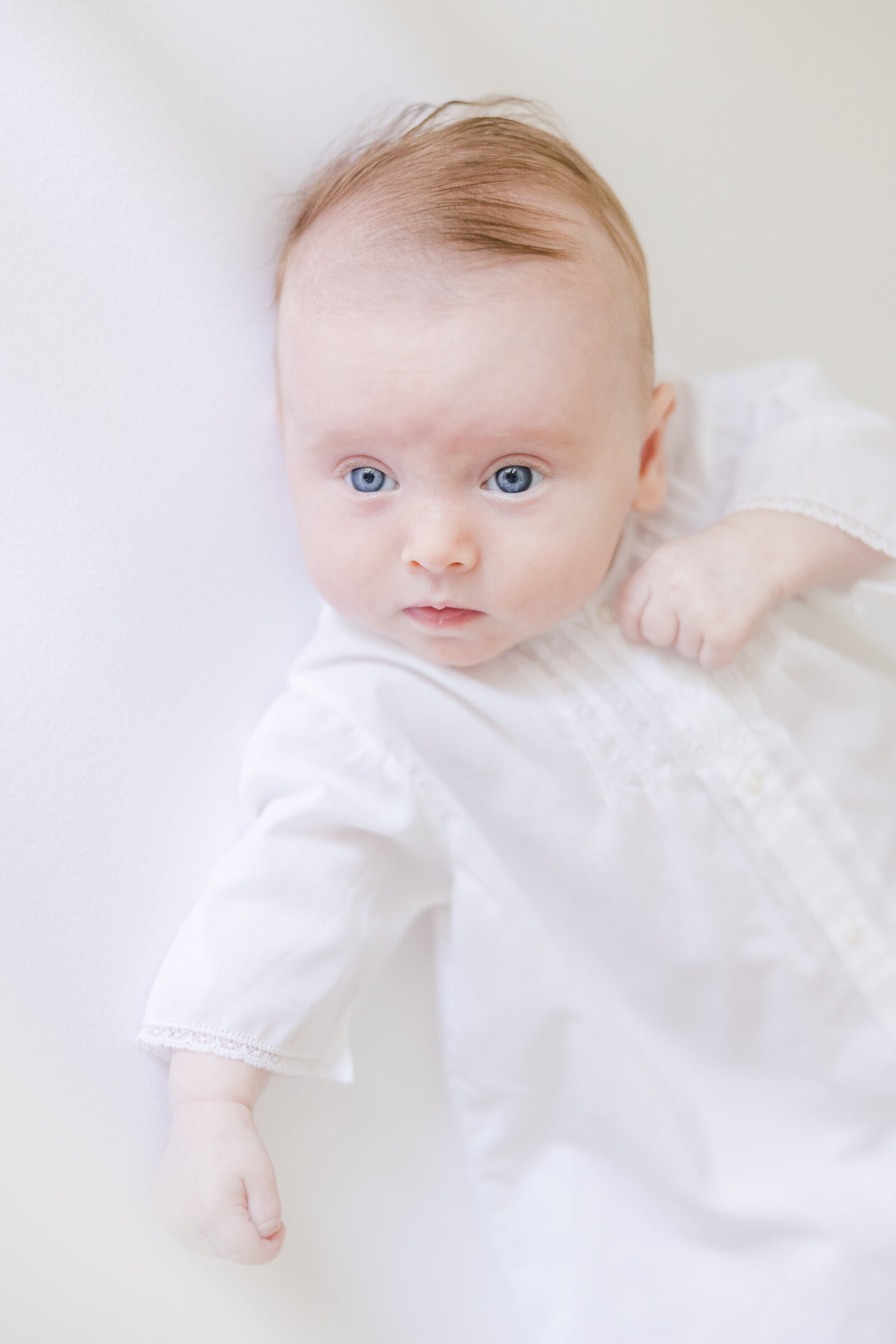 Blue eyed baby in white gown gazing at the camera.