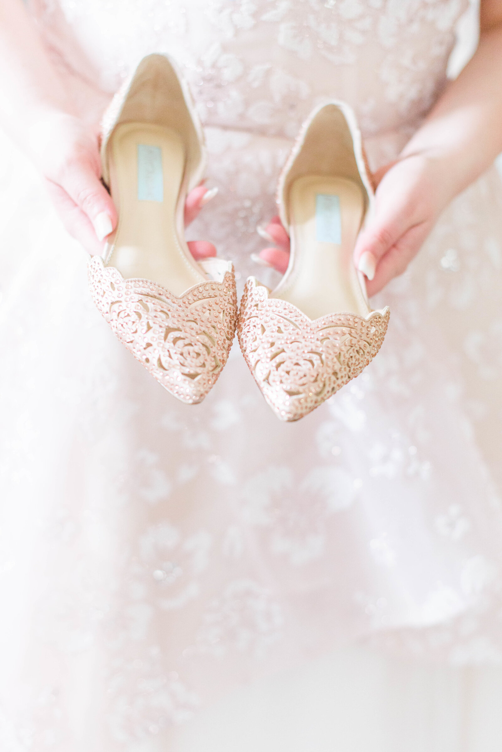 Bride holds blush shoes in front of blush wedding dress on wedding day photographed by Cait Kramer Photography
