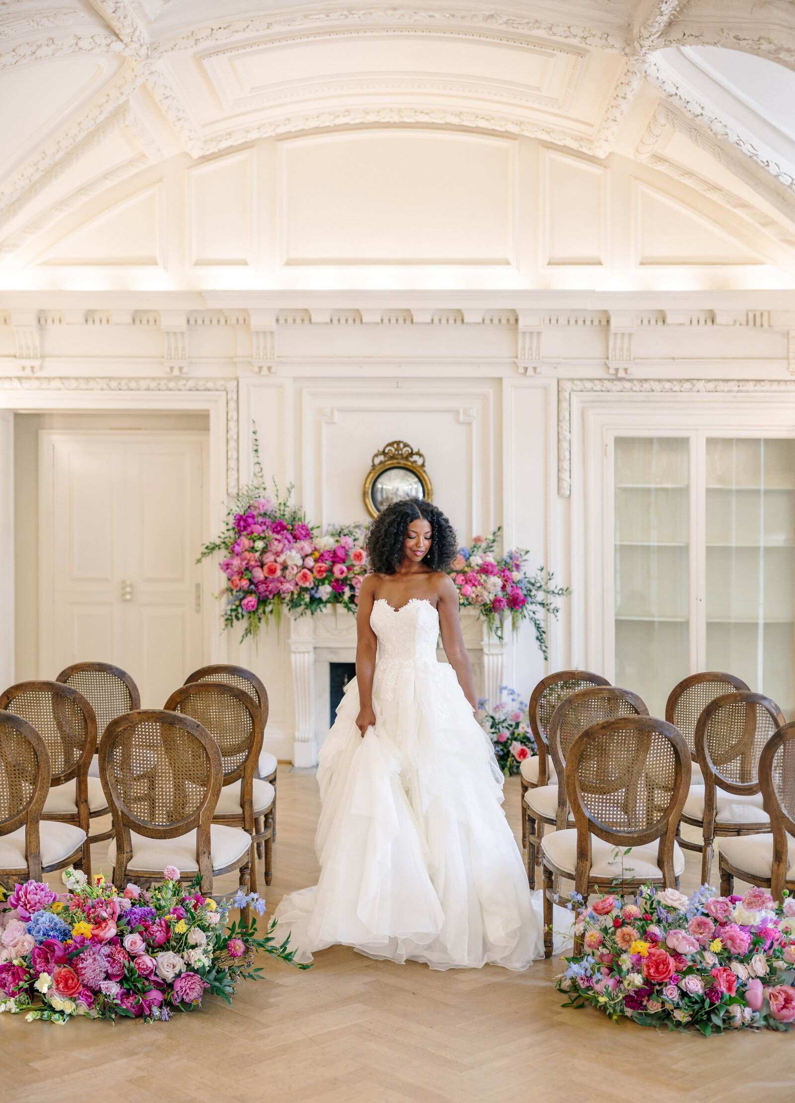 A bride in a white gown stands in an elegant, white room with a vaulted ceiling. She is holding her dress and is surrounded by wooden chairs, and vibrant floral arrangements in pink and purple hues.