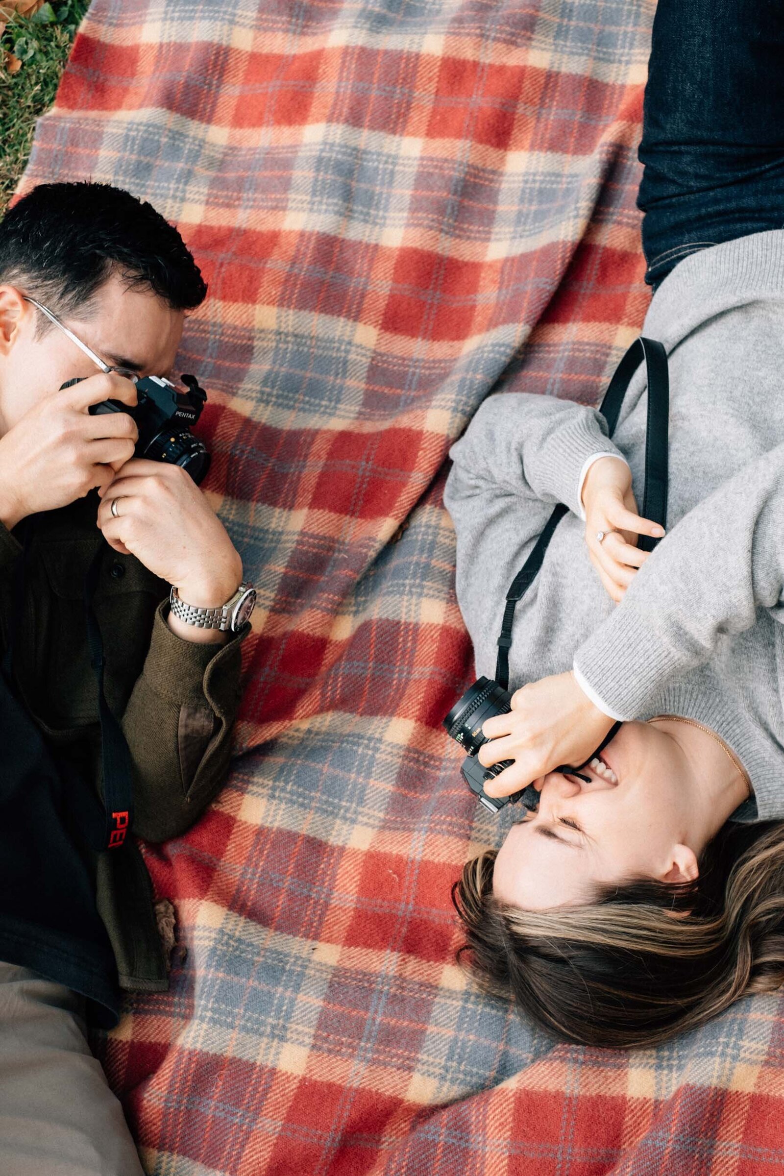 A couple lays on a plaid blanket and takes photos of each other with vintage film cameras during a park engagement shoot.