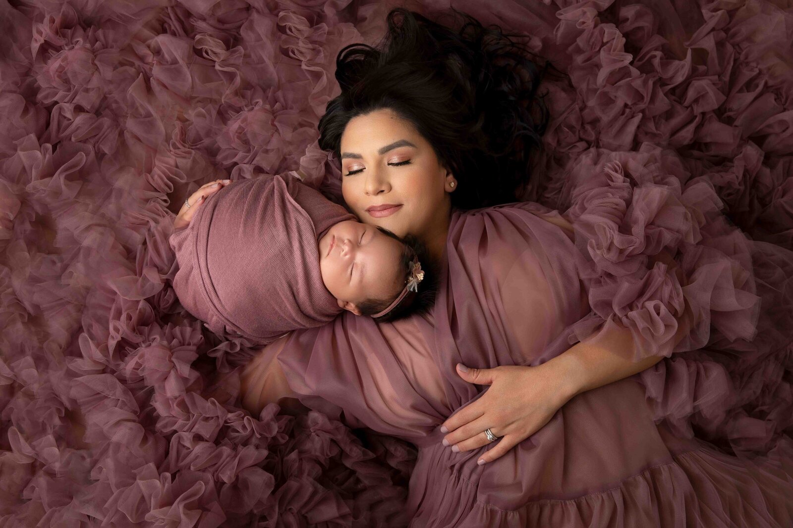 A loving mother cradles her newborn baby while posed gracefully on the floor, dressed in a stunning mauve fluffy couture gown that envelops both mom and baby. This elegant portrait, captured at Little Legacies Studio in Phoenix, Arizona, highlights the beauty of motherhood and the tender connection between mom and baby.
