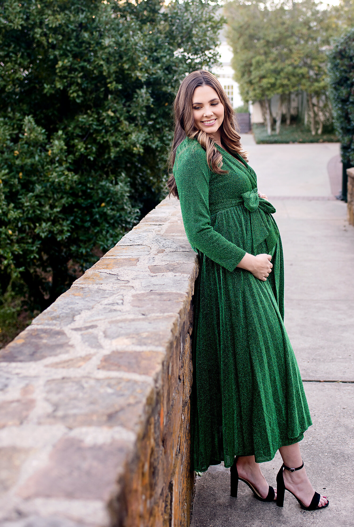 A pregnant woman wearing a green dress and black high heels standing on a stone bridge at her maternity photo shoot at Turtle Creek Park in Dallas.