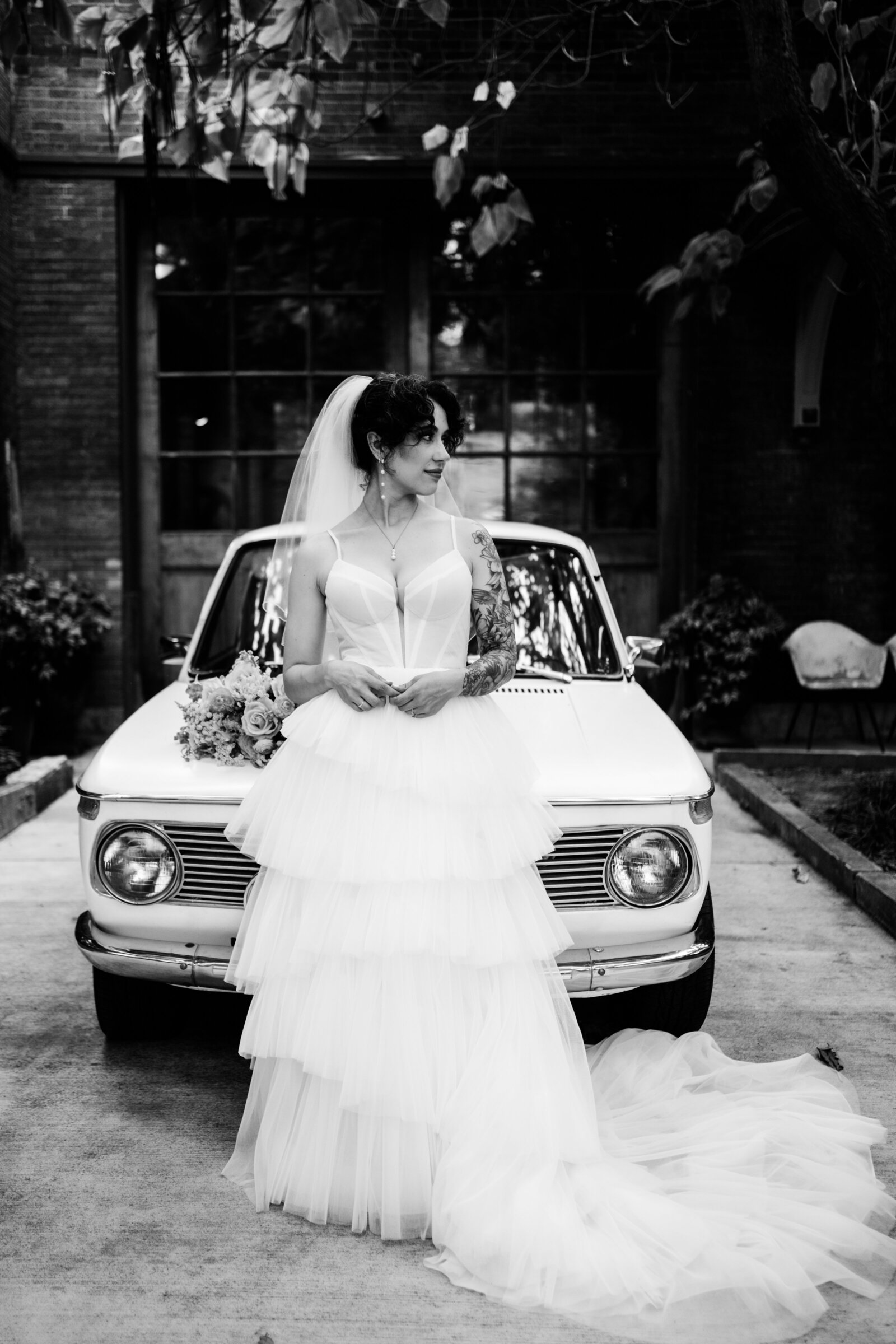 Bridal portrait in a downtown STL venue. She is posing in front of a vintage car holding her hands together and looking left. Her dress is layered and fitting.