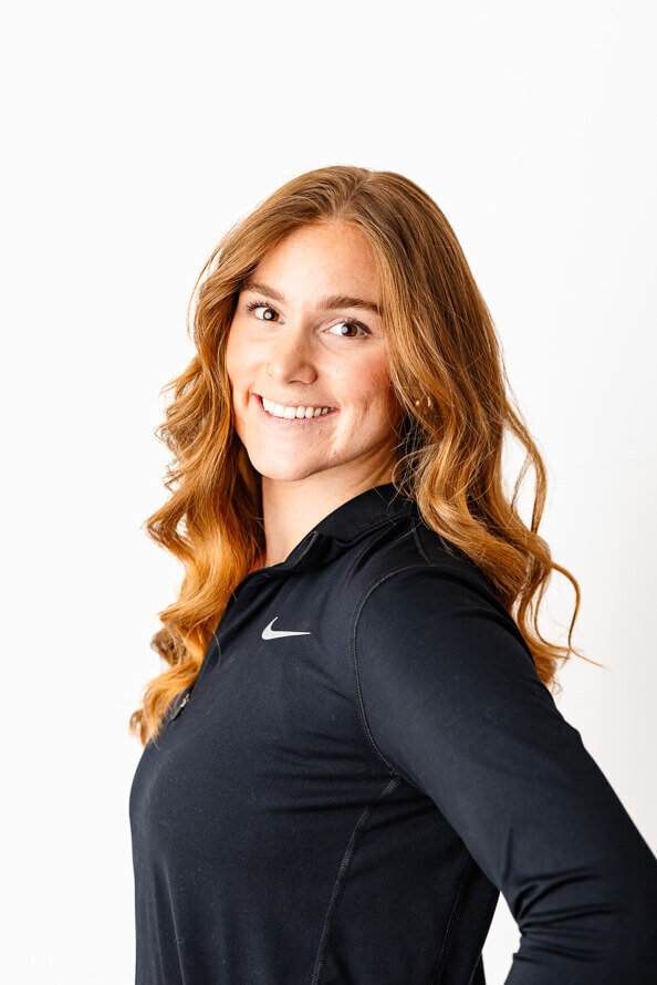 Woman in black athletic shirt and red hair smiles at camera in a white photo studio.