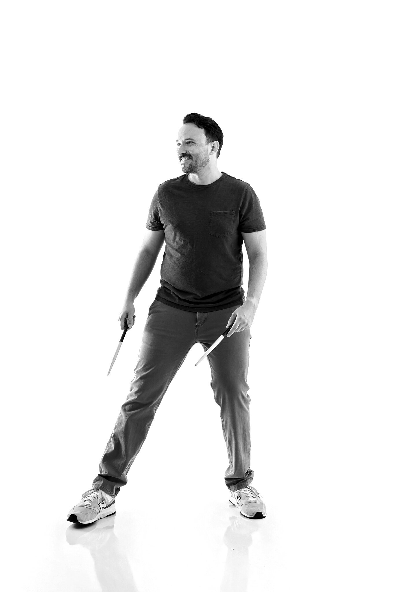A man stands smiling, holding drumsticks in both hands. Dressed casually, he poses against a white background in this relaxed black and white shot by a Jacksonville band photographer.