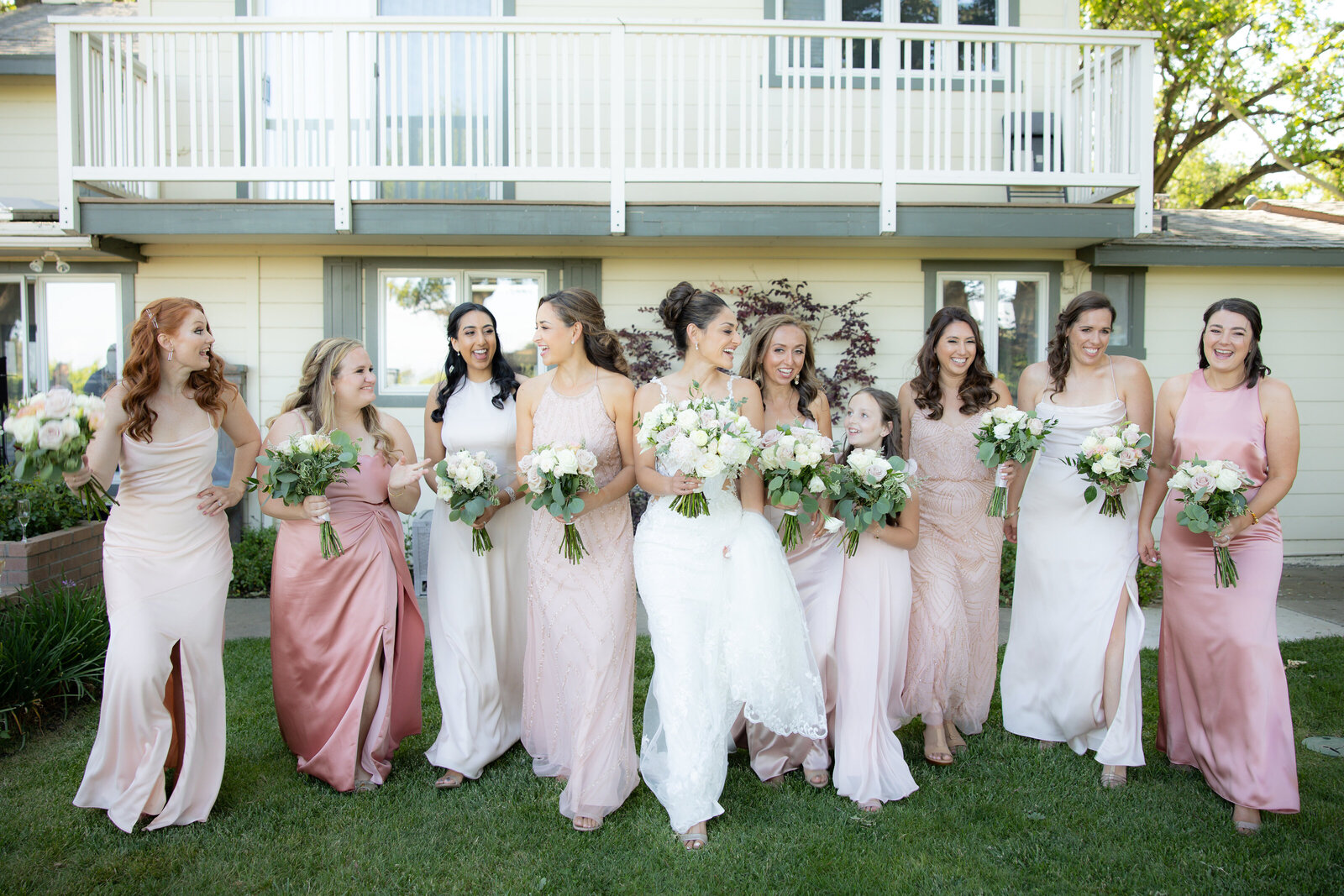 Bride walks with her bridesmaids who are dressed in different shades of pink.  They laugh and walk in front of a house. Photo captured by sacramento wedding photography studio, philippe studio pro.
