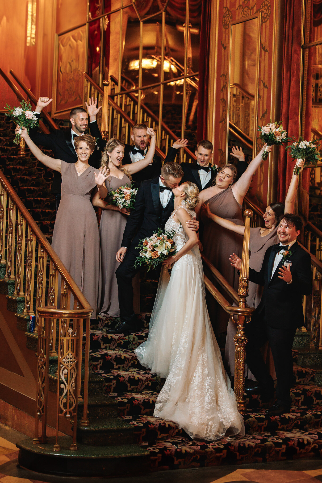 Positioned on a a grand staircase a newly wed couple shares a kiss surrounded by their bridal party cheering in excitement 