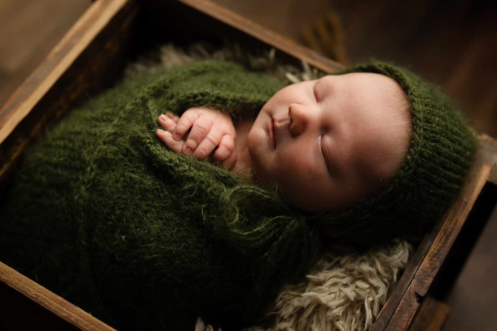 Newborn boy wrapped in an army green knit wrap and bonnet, posed in a rustic wooden crate