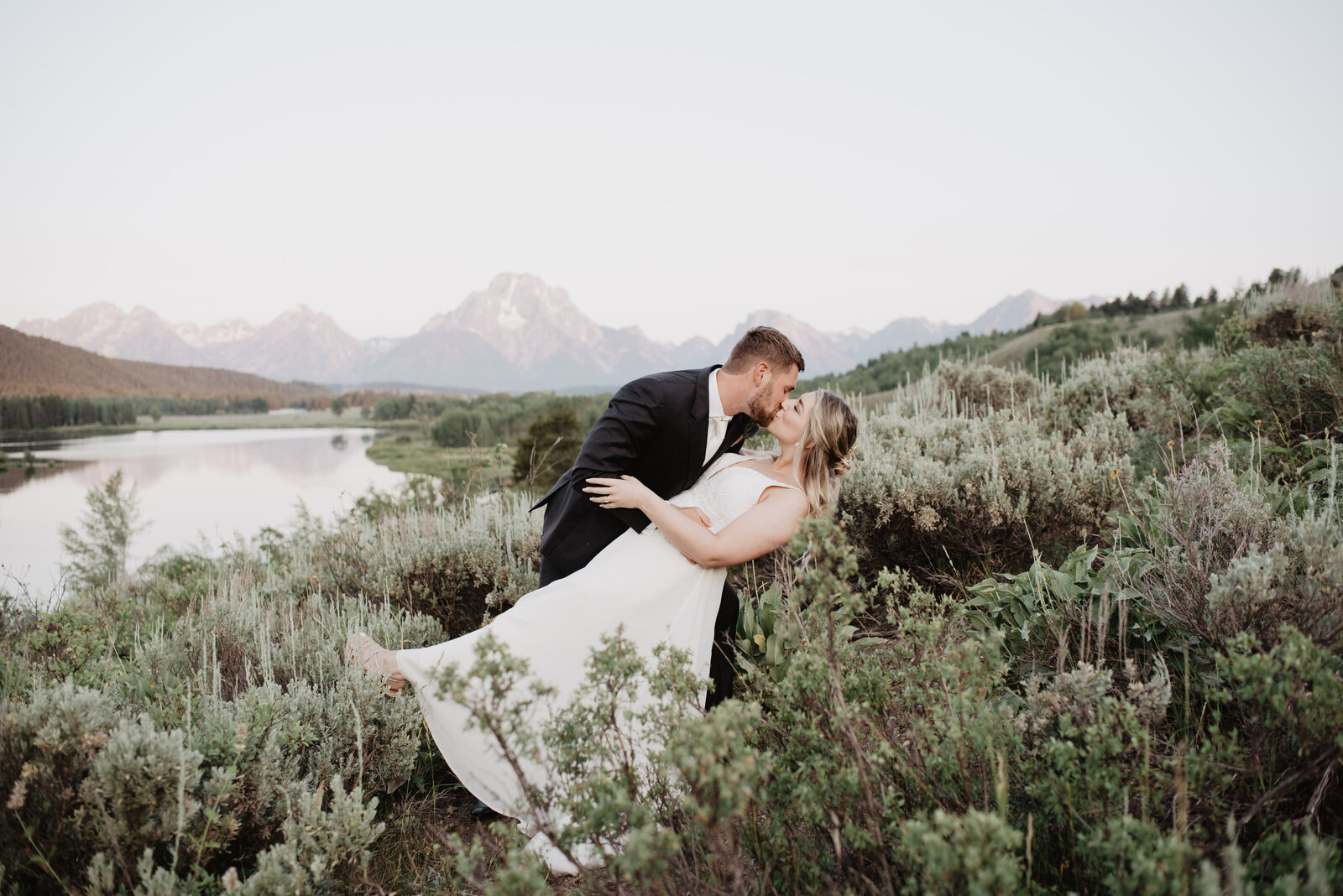 oxbow bend wedding with bride and groom kissing as they dance together in front of a river with the mountains in the far distance for their wyoming elopement