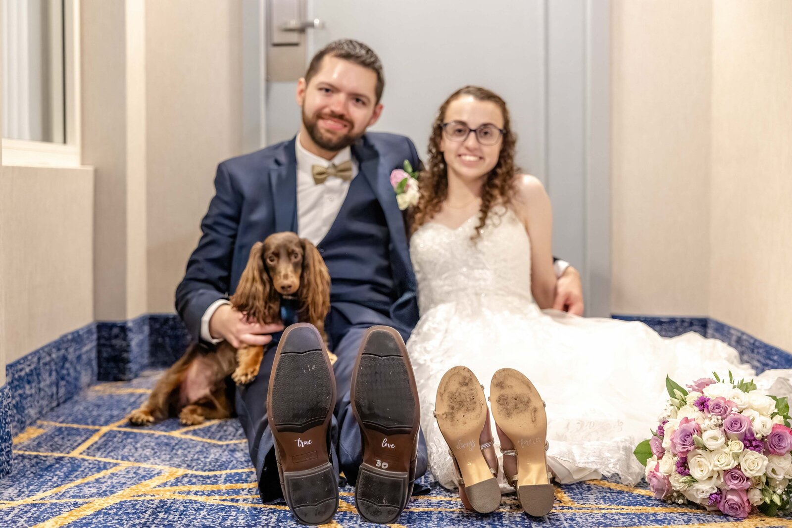 The bride and groom sit on the floor with their dog, holding hands, surrounded by wedding attire and flowers, captured during a Wilmington, DE wedding, showcasing authentic moments and pet-friendly charm.