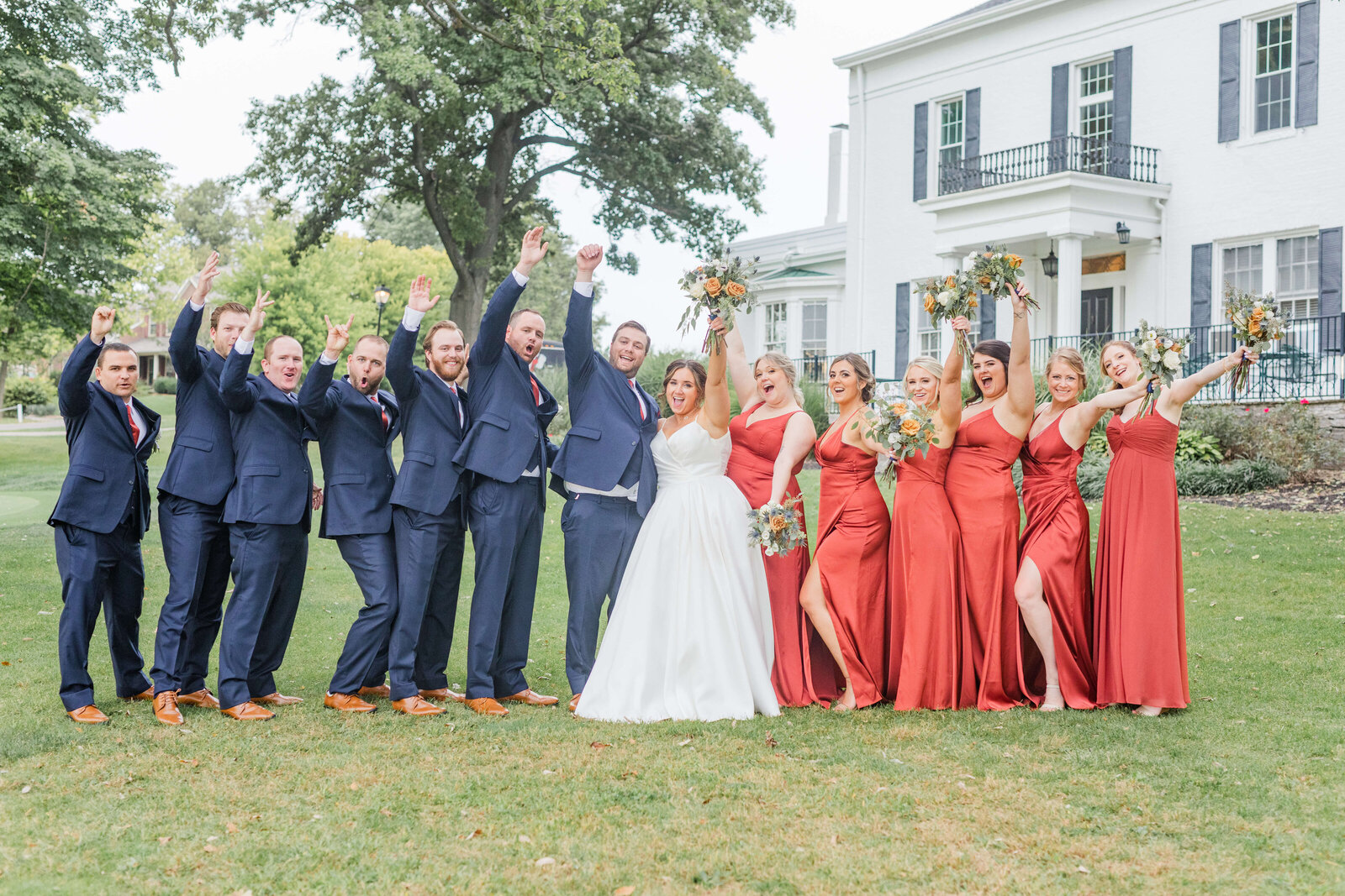 Bride and groom pose with their bridal party at their wedding and cheer with excitement. The bridesmaids are in orange and groomsmen in blue.