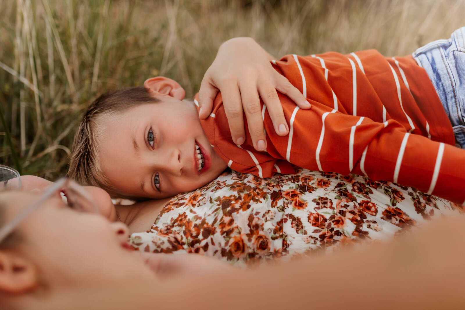 Overland Park Family Photographer captures mom in floral dress laying in field with son hugging on her chest