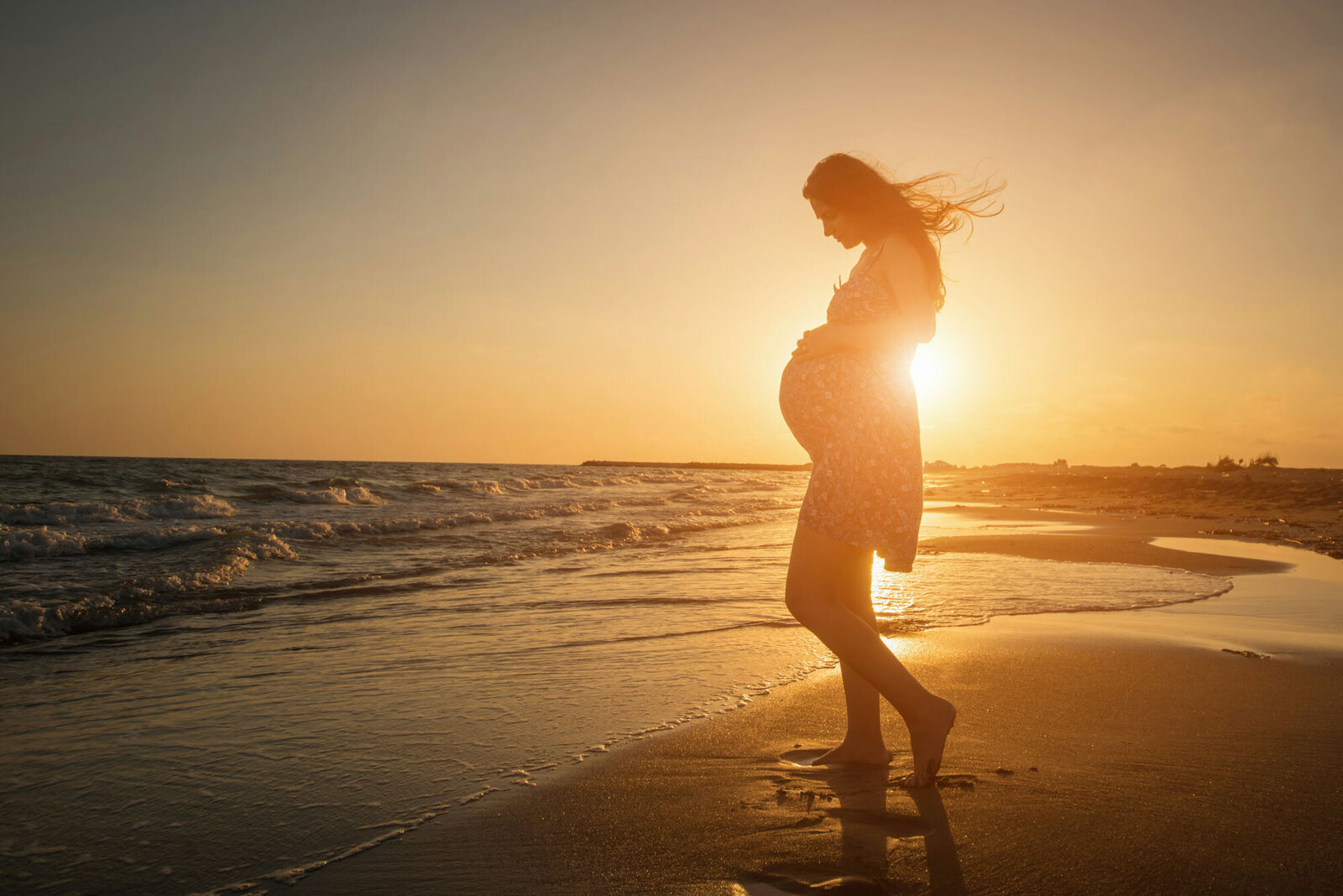 Pregnant woman is standing on the beach at sunset holding her belly