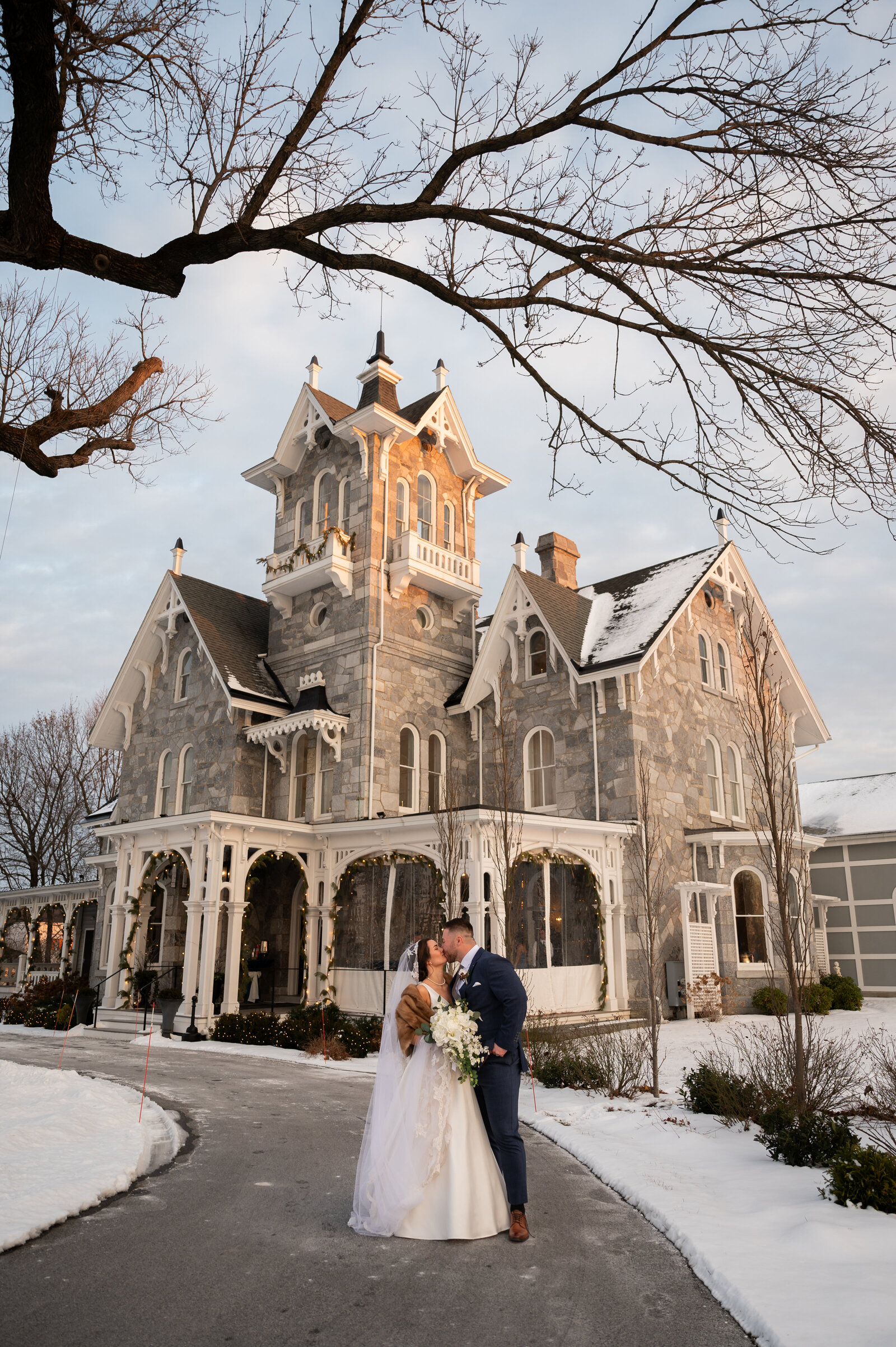 A bride and groom kiss outside a large, historic stone mansion in winter, with snow covering the ground and bare trees in the background.