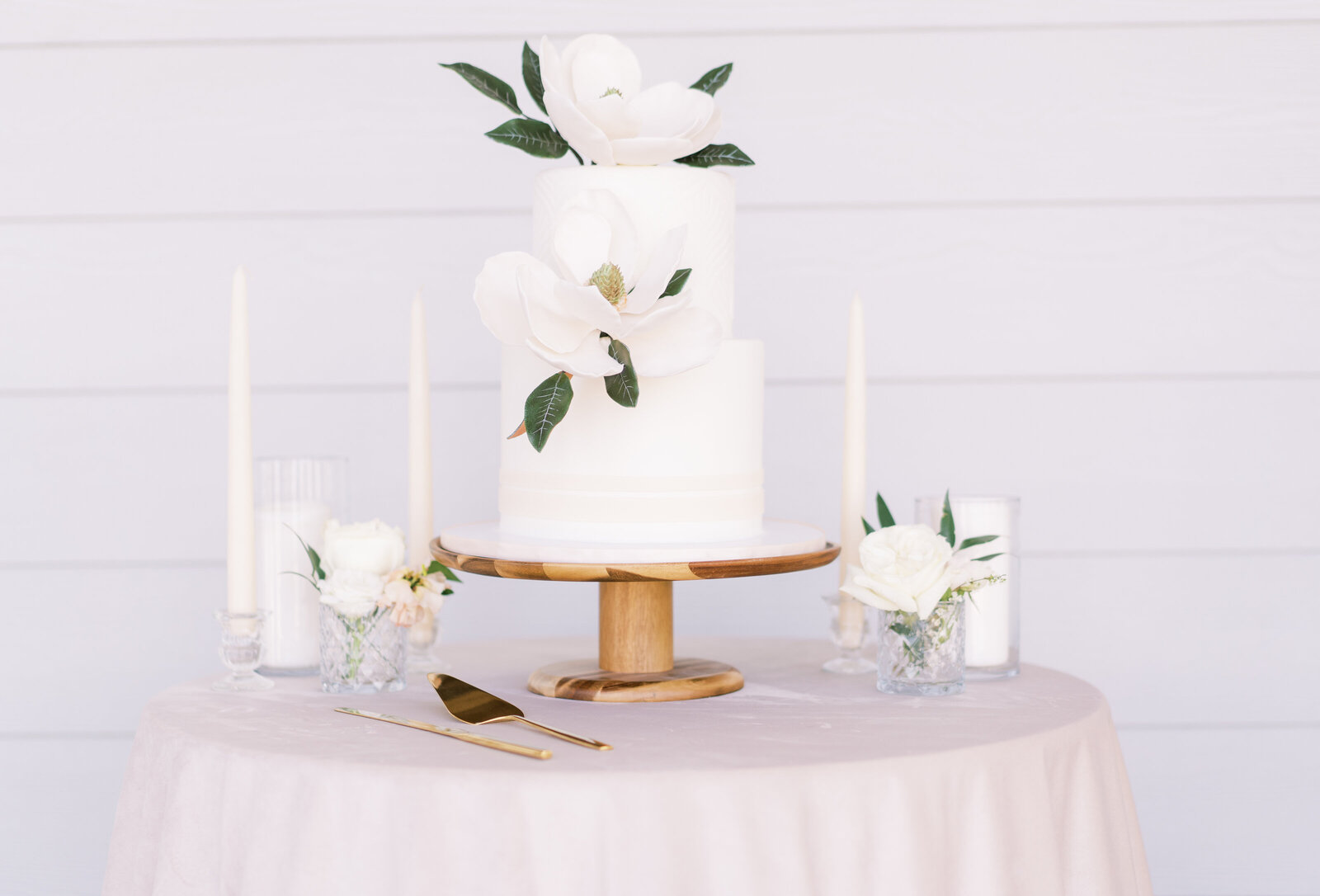 Portrait of two-tiered white wedding cake with flowers, candlesticks, and glassware atop a round table with blush tablecloth.