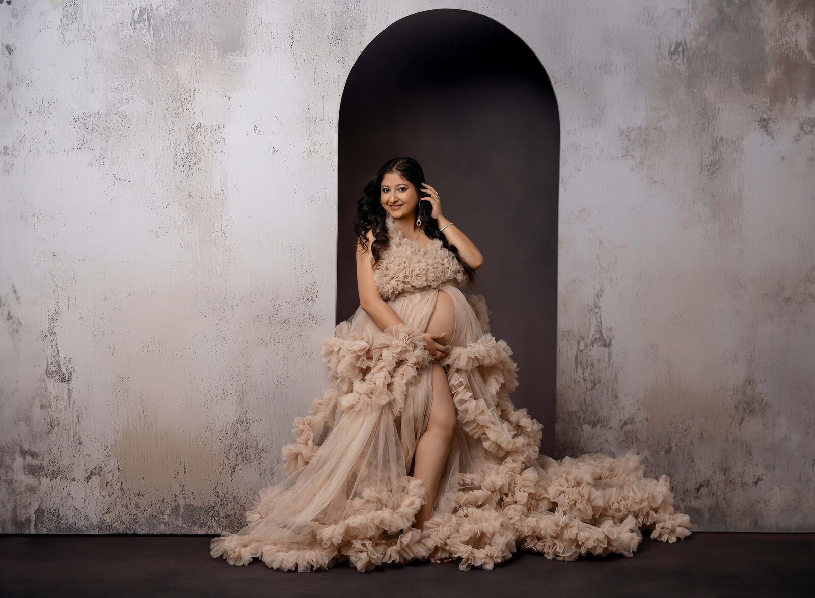 Pregnant women in cream ruffled maternity gown, holding belly and smiling at camera, in front of large arch wall. Photo taken by Jenni Nikki at Maternity photo studio in Atlanta, GA.