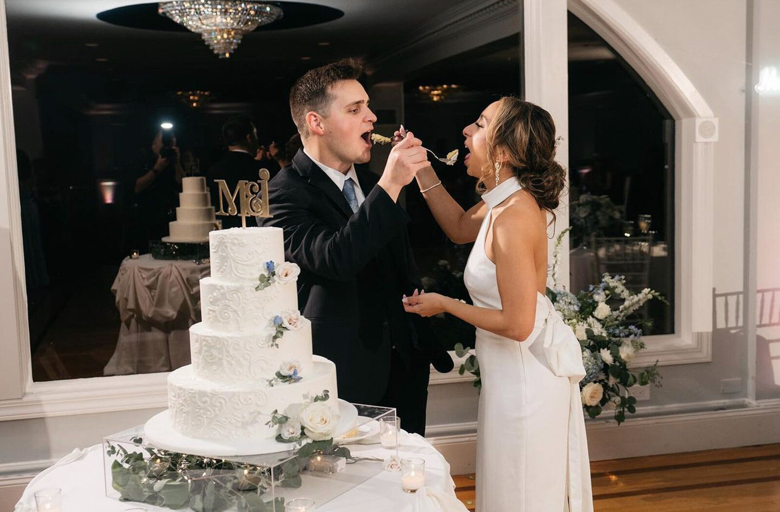 ewlywed couple stands by a four-tiered white wedding cake, feeding each other a slice