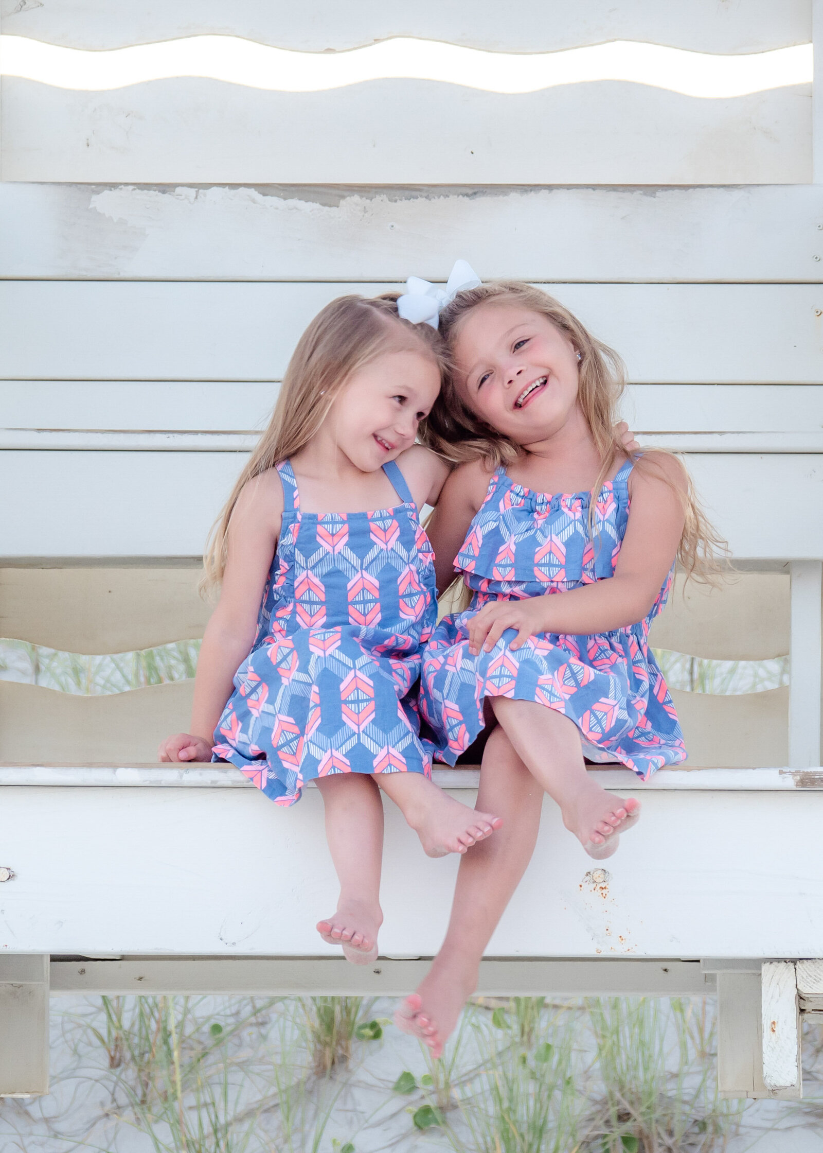 Two young sisters sit on a beach bench, wearing matching blue dresses with pink patterns and a white bow. Their joyful expressions capture a playful, sunny day by the sea, perfect for North Carolina family photography, highlighting sibling bonds.