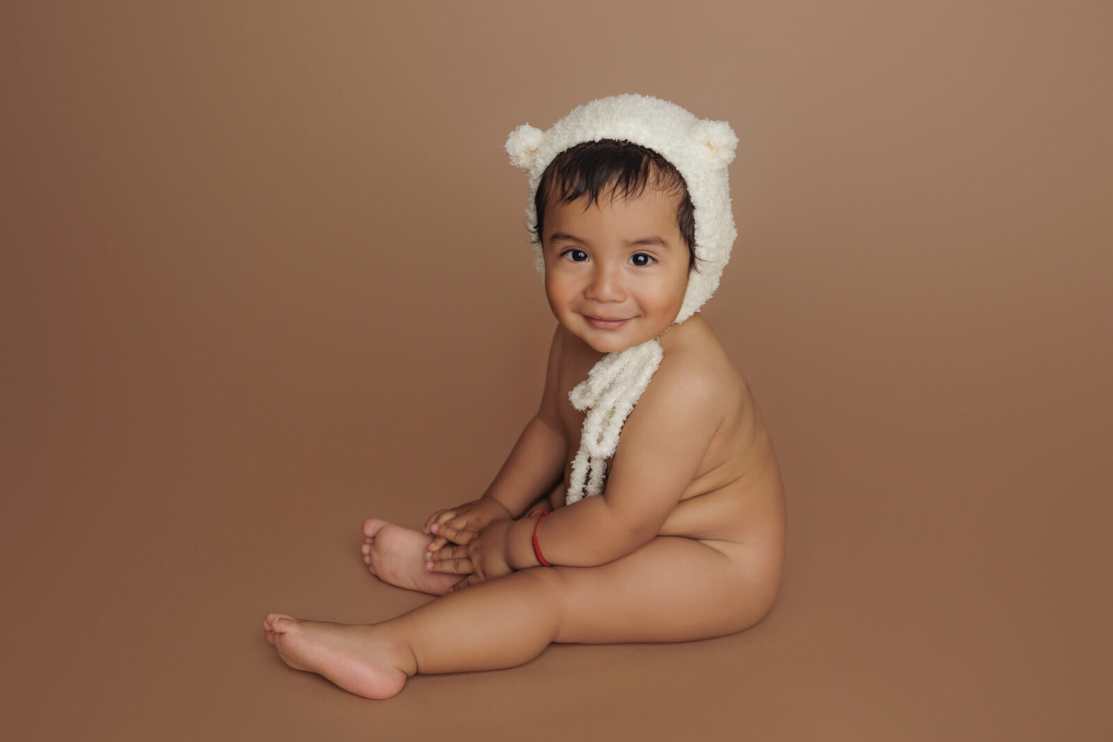 A cheerful baby with dark hair and bright eyes sits on a brown backdrop, wearing a white knit hat with small bear ears. The baby is naked, smiling warmly at the camera, with a red string bracelet on their wrist, creating a cozy and adorable portrait.