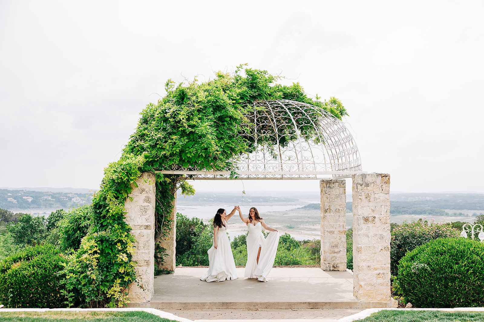Two women in white dresses joyfully dance together under a vine-covered stone gazebo, scenic hills and water in the background—perfect inspiration from a San Antonio Wedding Photographer.