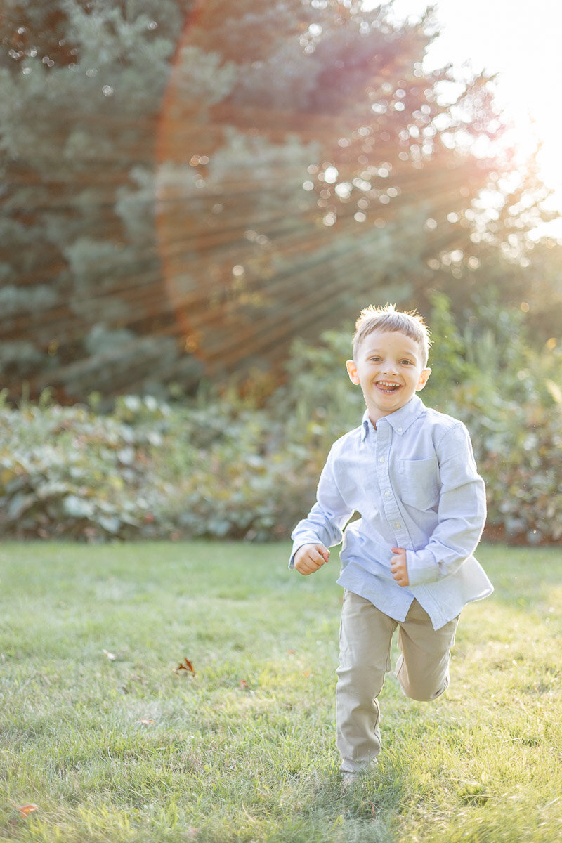 Young boy running joyfully across a sunlit field, beaming with a big smile as golden rays shine through the trees behind him.