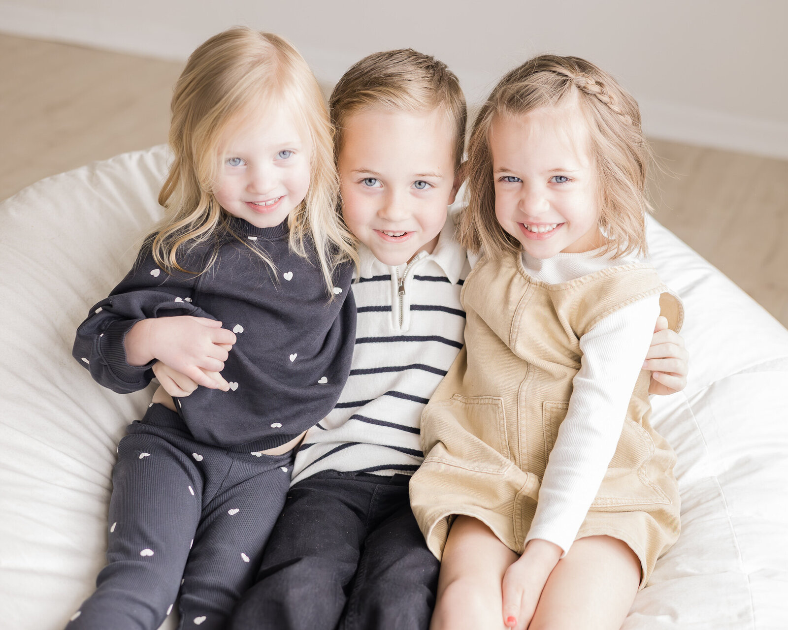 Three siblings sitting on a bean bag smiling at the camera.  Brother has sandy blonde hair wearing a white and black striped shirt, black pants, and smiling at the camera.  Youngest sister has blone hair wearing a blue and white dotted long sleeve shirt and pants, smiling away from the camera.  Other sister has sandy blonde hair, wearing a light tan dress, with a white shirt under it, smiling at the camera.