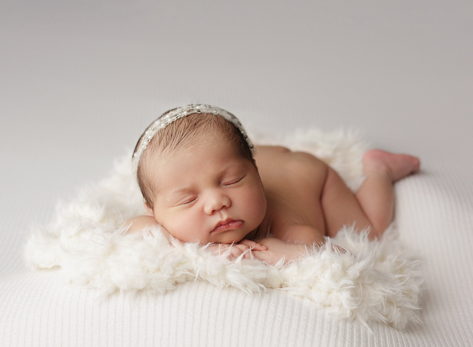 A precious newborn baby sleeps peacefully on their stomach, head resting on hands adorned with a delicate headband, surrounded by a fluffy white blanket. Captured by an Orange County newborn photographer, this serene moment is a testament to the artistry of baby photography.