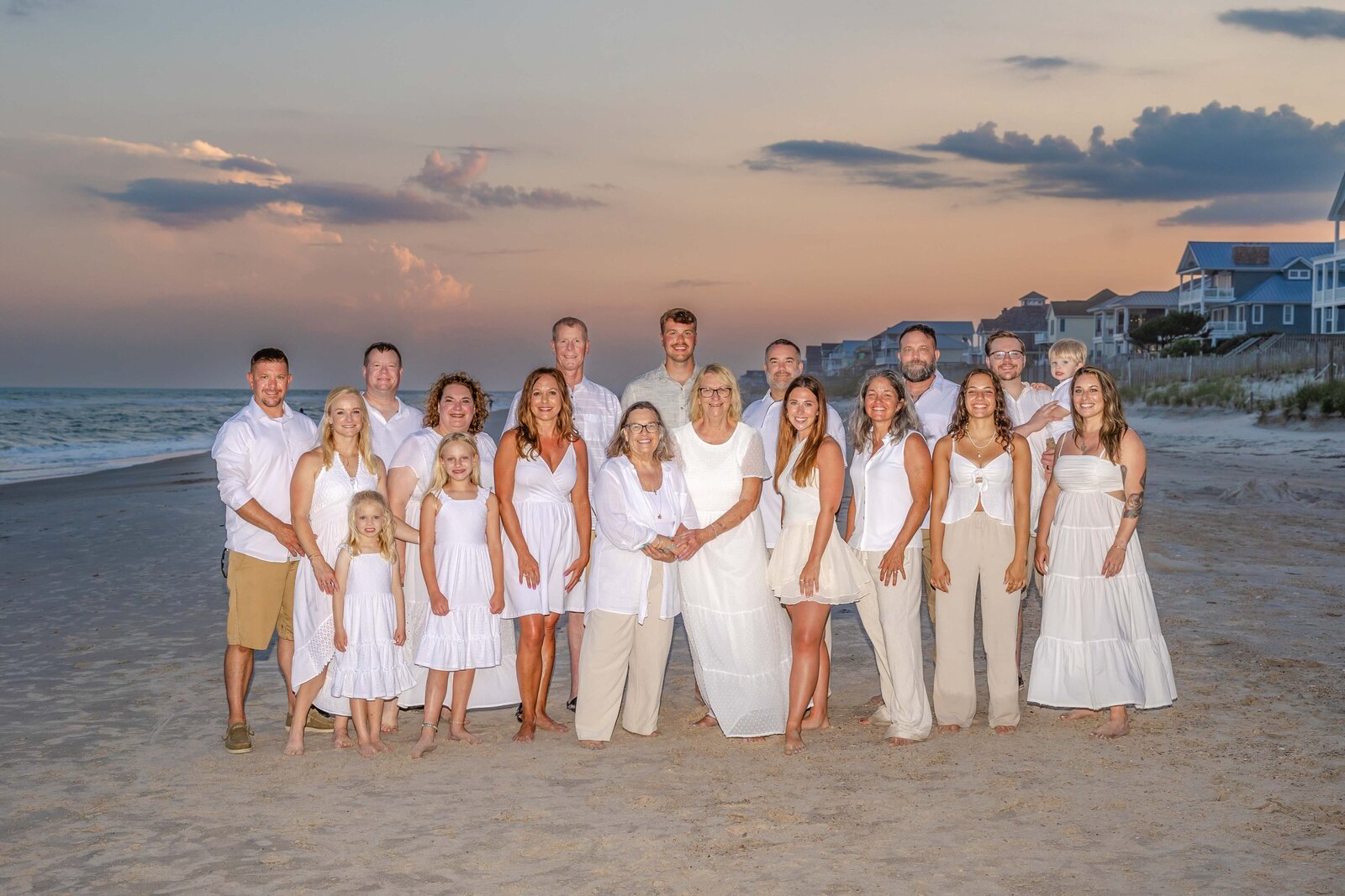 A large family dressed in white poses on the beach at sunset, with soft pastel skies and beachfront homes in the background. This image captures the elegance and warmth of a family gathering, perfect for artistic family photography in Wilmington, NC.