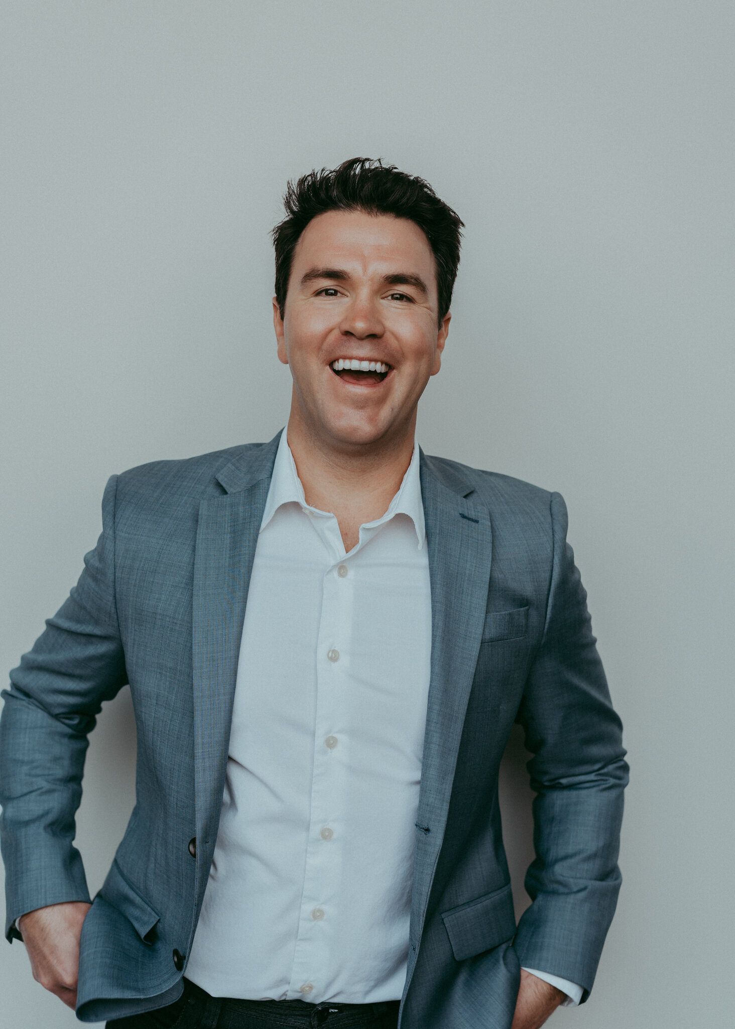 A male realtor shows off his fun and welcoming personality during his personal branding session. He is wearing a white button up shirt, dark denim jeans, and a slate blazer as he makes iContact with an open mouth smile.